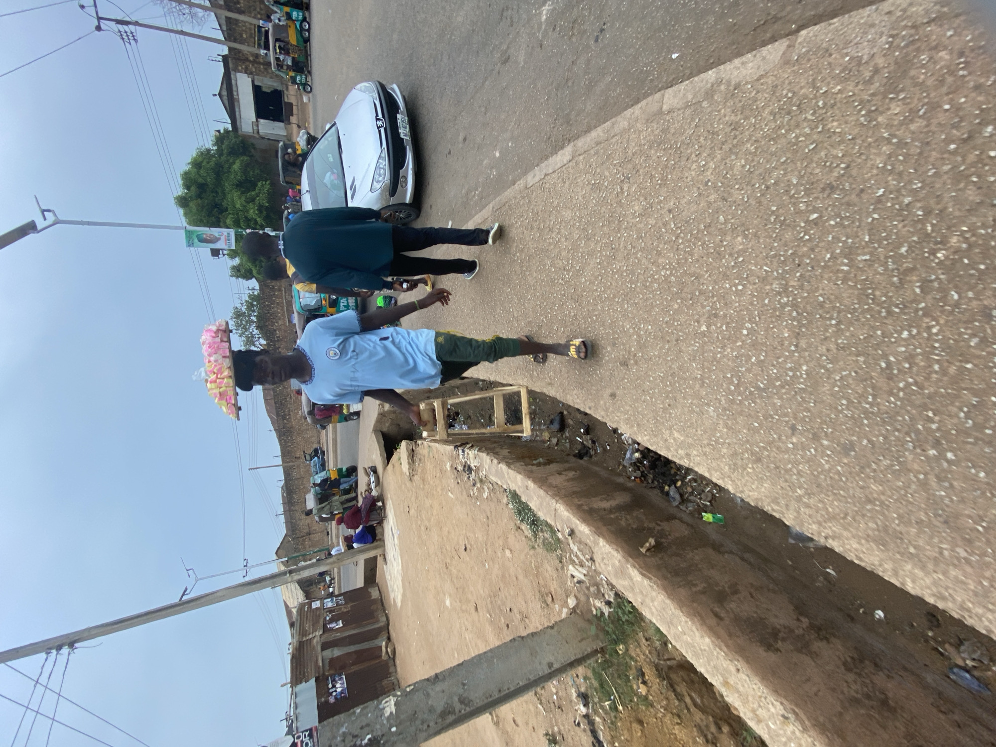 This image captures a vibrant street scene in Bukuru, Nigeria, during what appears to be daytime under a clear, bright sky. The perspective is slightly elevated, looking down along a road and an adjacent raised concrete pathway or barrier.
In the foreground, a young man, dressed in a light blue t-shirt bearing a "MAN CITY" logo and dark shorts, walks barefoot on the concrete barrier, balancing a tray of colorful, wrapped goods (possibly snacks or sweets) on his head. He wears yellow flip-flops. To his right, another person, seen from behind in a dark blue or green top and black trousers, walks on the asphalt road.
A silver Peugeot car (possibly a 206 model) is parked or moving slowly on the paved road. Its license plate has partially visible characters, starting with "KJM" or "KJN." In the background, typical Nigerian commercial tricycles, also known as Keke Napep, in various colors like green and yellow, are visible, suggesting a busy thoroughfare or market area. One tricycle shows "PTA" text. Several other individuals are scattered in the mid-ground and background, some seated or standing near the tricycles and buildings.
The street is lined with simple, low-rise buildings, some featuring corrugated iron facades or fencing. Utility poles with overhead power lines stretch across the scene, and a street lamp pole is also visible. A large, lush green tree stands prominently in the mid-ground. The area between the concrete barrier and the road appears to be a drainage ditch containing visible litter and dirt. A small, simple wooden structure is also present near the barrier. A distant banner on a pole displays a person's face, though the text is unreadable.