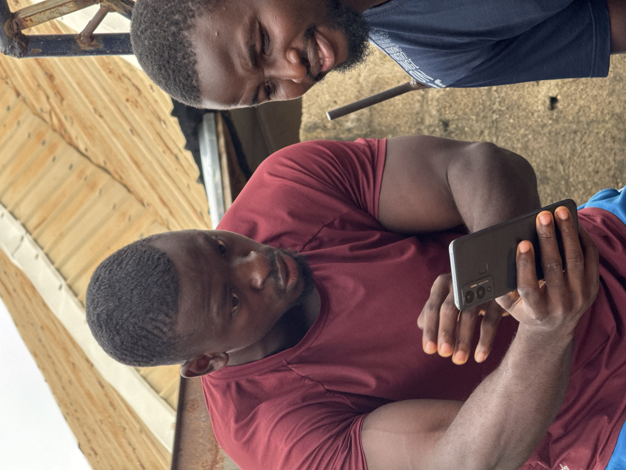Two young Black men are looking at a smartphone held by the man on the left. The man on the left is wearing a maroon t-shirt and has dark skin and short, dark, wavy hair. He is holding the smartphone with both hands, his fingers are visible and are dyed orange. The man on the right is wearing a dark blue t-shirt and has dark skin, a beard, and short, dark hair. He is smiling. They are standing outdoors, with a corrugated metal roof and a textured wall in the background. The scene appears to be in an urban or semi-urban setting in Akwanga, Nigeria, possibly a residential area or a local business.