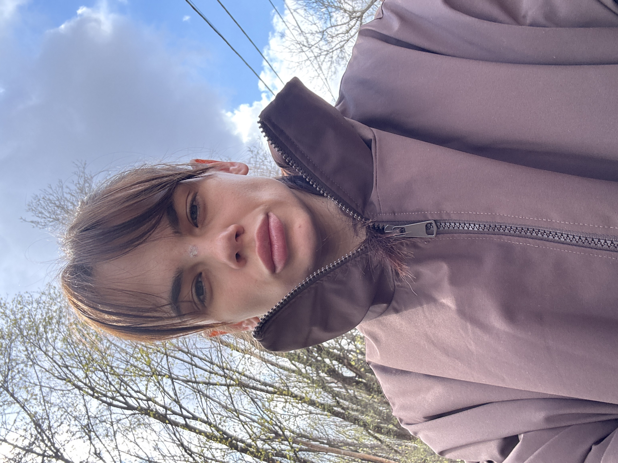 A young woman is taking a selfie outdoors, with the camera pointed upwards at her face. She has brown hair with bangs and light skin. She is wearing a brown jacket with a zipper that is partially closed. Behind her, bare tree branches are visible against a cloudy sky with power lines. The exact location within the city of Rostov-on-Don, Russia is not identifiable from the image.