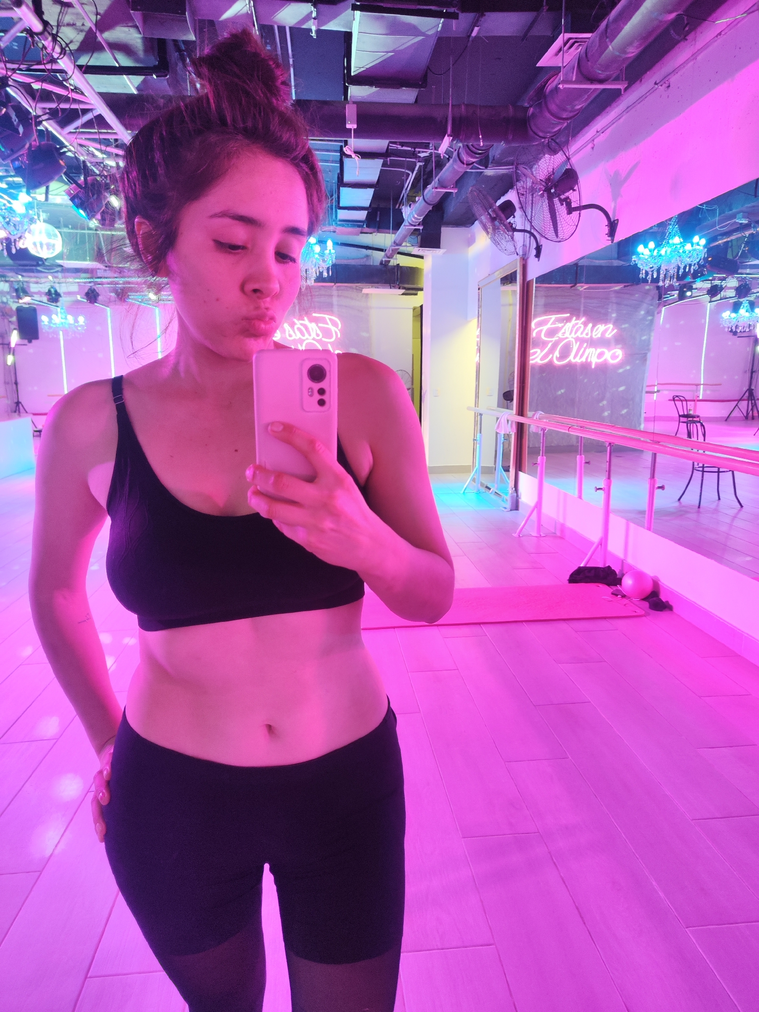 A young woman with her hair in a bun is taking a mirror selfie in a dance studio. She is wearing a black sports bra and black leggings. The studio is lit with pink and purple lights, with disco balls and chandeliers visible in the background. A neon sign on the mirror reads "Estás en el Olimpo". The floor is made of light-colored wood planks. The image was taken in Santiago, Chile.