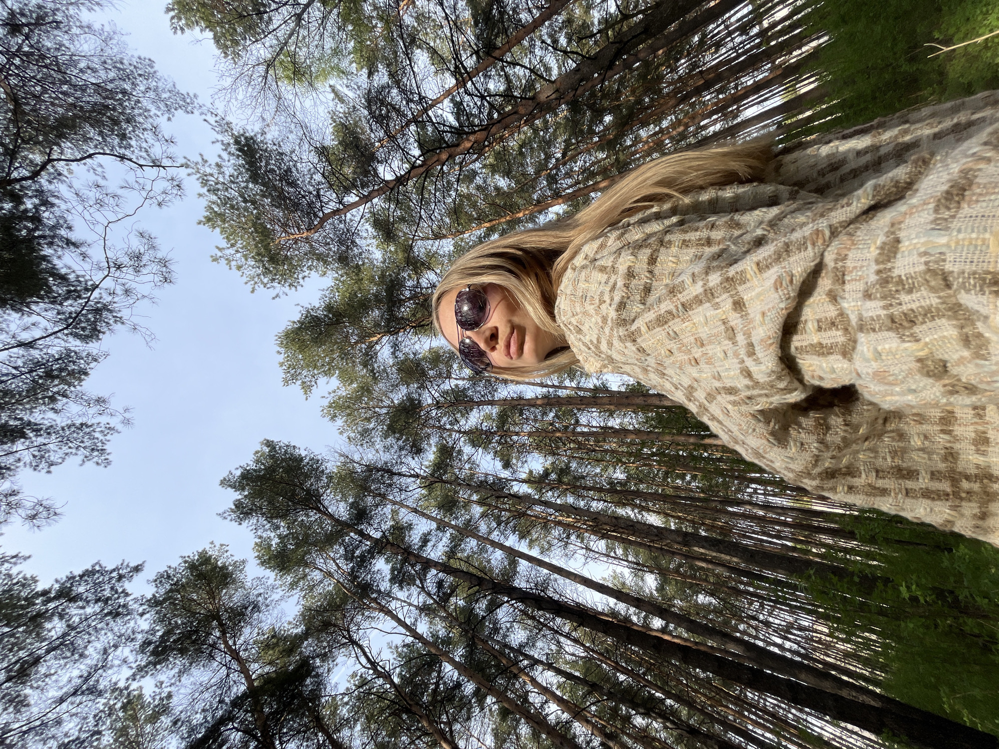 A low-angle shot shows a young woman in sunglasses and a tweed-like jacket, positioned against a backdrop of tall trees and sky. The trees are slender and numerous, reaching upwards, suggesting a dense forest. The sky is pale blue and visible through the spaces between the branches. The woman is wearing a jacket and sunglasses. The environment is a forest. The exact location in the city of Chaikovsky, Russia is not discernable from the provided image.