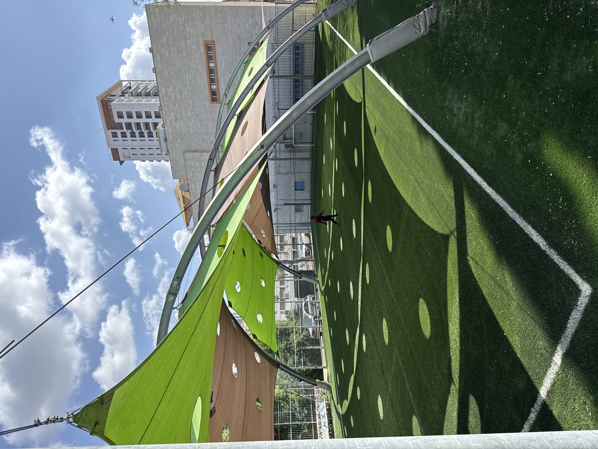 The image shows an outdoor area in Petah Tikva, Israel. There is a green artificial turf field with white lines. A person in red clothing is standing in the center of the field. Overhead, a metal frame supports a shade structure made of green and brown material with circular cutouts. Shadows from the shade structure create a pattern of circles on the field. In the background, there are buildings under a partly cloudy sky.
