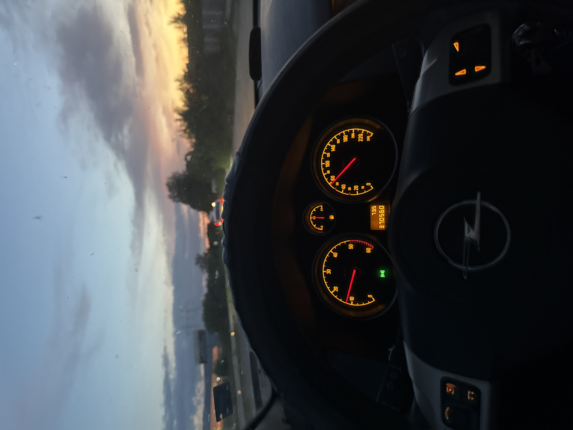 The image depicts the interior of a car, specifically the dashboard and steering wheel, with an Opel logo prominently displayed. The instrument panel is illuminated, showing the speedometer, fuel gauge, and odometer reading 370560. Through the windshield, a view of the outside is visible, featuring a sky with twilight colors and trees lining a road. The image was taken in the city of Plovdiv, Bulgaria.