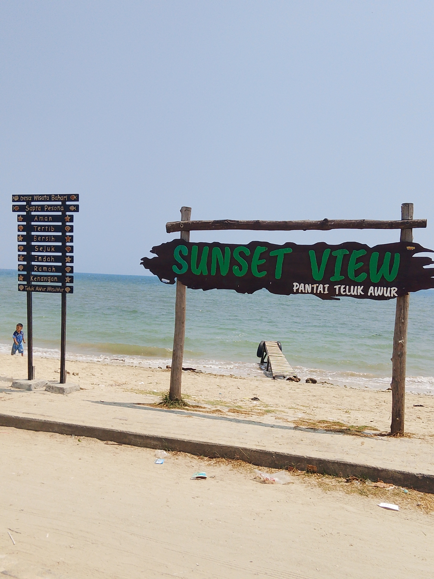 The image shows a beachfront scene in Pejagan, Indonesia. A wooden sign with "SUNSET VIEW" painted in green is prominently displayed, with smaller text below reading "PANTAI TELUK AWUR". To the left, a black sign with multiple rows lists various amenities and attributes, starting with "Desa Wisata Bahari". A young boy in a blue and white shirt and shorts stands on the sandy beach near the sign. In the background, the ocean stretches out under a clear sky, with gentle waves reaching the shore. A small wooden boardwalk leads from the beach into the water. The overall environment is that of a coastal tourist area.