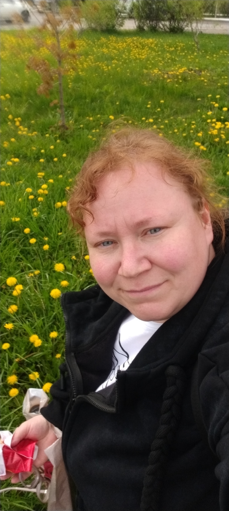 The image shows a woman in an outdoor setting, specifically a field of dandelions in Chaikovsky, Russia. The woman has reddish-blonde hair and blue eyes, and she is wearing a black hooded jacket over a white shirt. She is holding an object wrapped in red and white paper in her hand. The background features a green field dotted with yellow dandelions. A small tree is visible in the background, as well as glimpses of a vehicle and other greenery, possibly bushes or trees. The overall scene suggests a casual outdoor setting in a park or natural area within the city.
