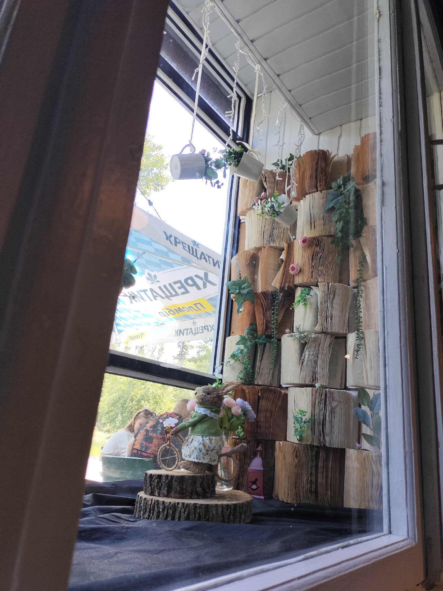 A store display is visible through a window in Unknown, Unknown. Inside, there is a small display featuring a layered, decorative arrangement of books, some with plants interspersed between them. Hanging above are decorative white cups, also containing small plants. In front of the book arrangement, there's a layered wood stump display. A small, dressed-up figure of a mouse with flowers and a miniature bicycle stands atop the display. A table with a black cloth forms the base of the display. Through the window, a sign with "ХРЕЩАТИ" is visible along with some trees. The angle suggests a view from inside a building, with a curtain partially obscuring the scene to the left. Two people can be seen through the window as well.