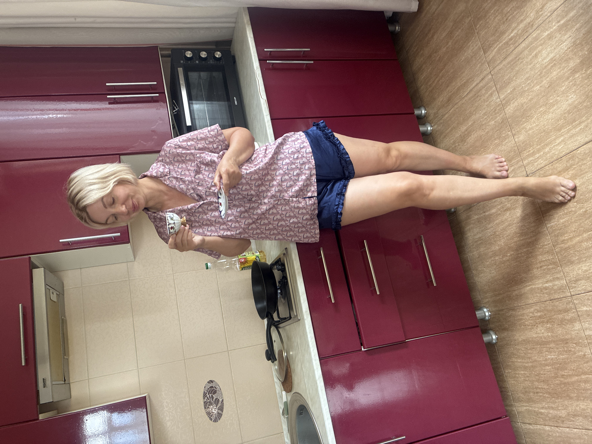 In a domestic kitchen, a blonde woman with short hair stands barefoot, holding a small cup and saucer. She wears a pink pajama top with a patterned design and navy blue ruffled shorts. She is looking down at the cup and saucer. Behind her, a black oven is visible, and to her left, a stovetop with a frying pan is present. The kitchen features dark red cabinets and light-colored tiled walls and flooring. The scene appears to be an interior shot of a home kitchen.