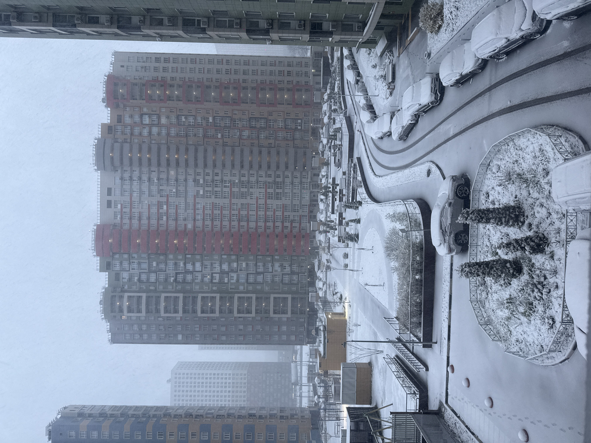 A high-angle shot, likely taken from an apartment window, depicts a snowy scene in Khimki, Russia. High-rise apartment buildings dominate the left side of the frame, with varying colors and architectural styles, partially obscured by a heavy snowfall that creates a hazy atmosphere.
To the right, a snow-covered parking area is visible, with numerous cars parked in rows along a curved road, most vehicles heavily laden with snow. Small circular garden plots dotted with snow-covered shrubbery punctuate the landscape. A pedestrian path runs parallel to the buildings, marked by footprints in the snow, suggesting recent activity. The overall scene conveys a wintery, urban landscape in a residential area.