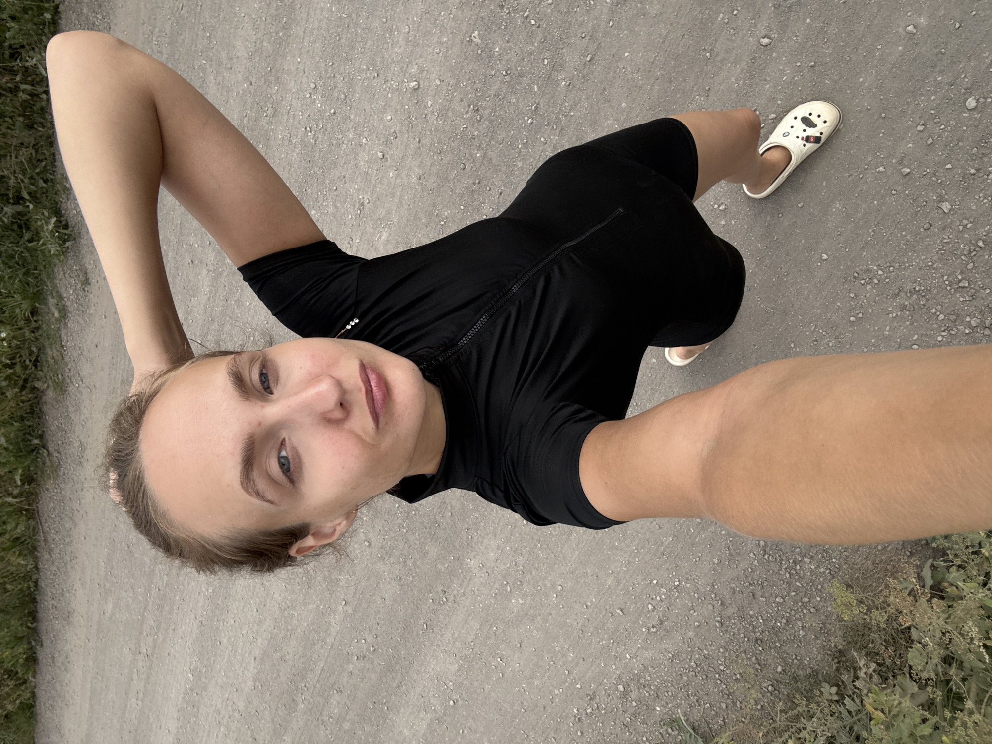 A young woman is taking a selfie outdoors, holding her right arm above her head. She is wearing a black short-sleeved athletic top with a zipper and black shorts. She has light-colored skin, light brown hair pulled back, and is wearing white clogs with cartoonish designs. The background is a dirt or gravel path with some dry grass and vegetation to the left. The scene appears to be in a rural or undeveloped area, consistent with the outskirts of a village like Podshivalovo, Russia.