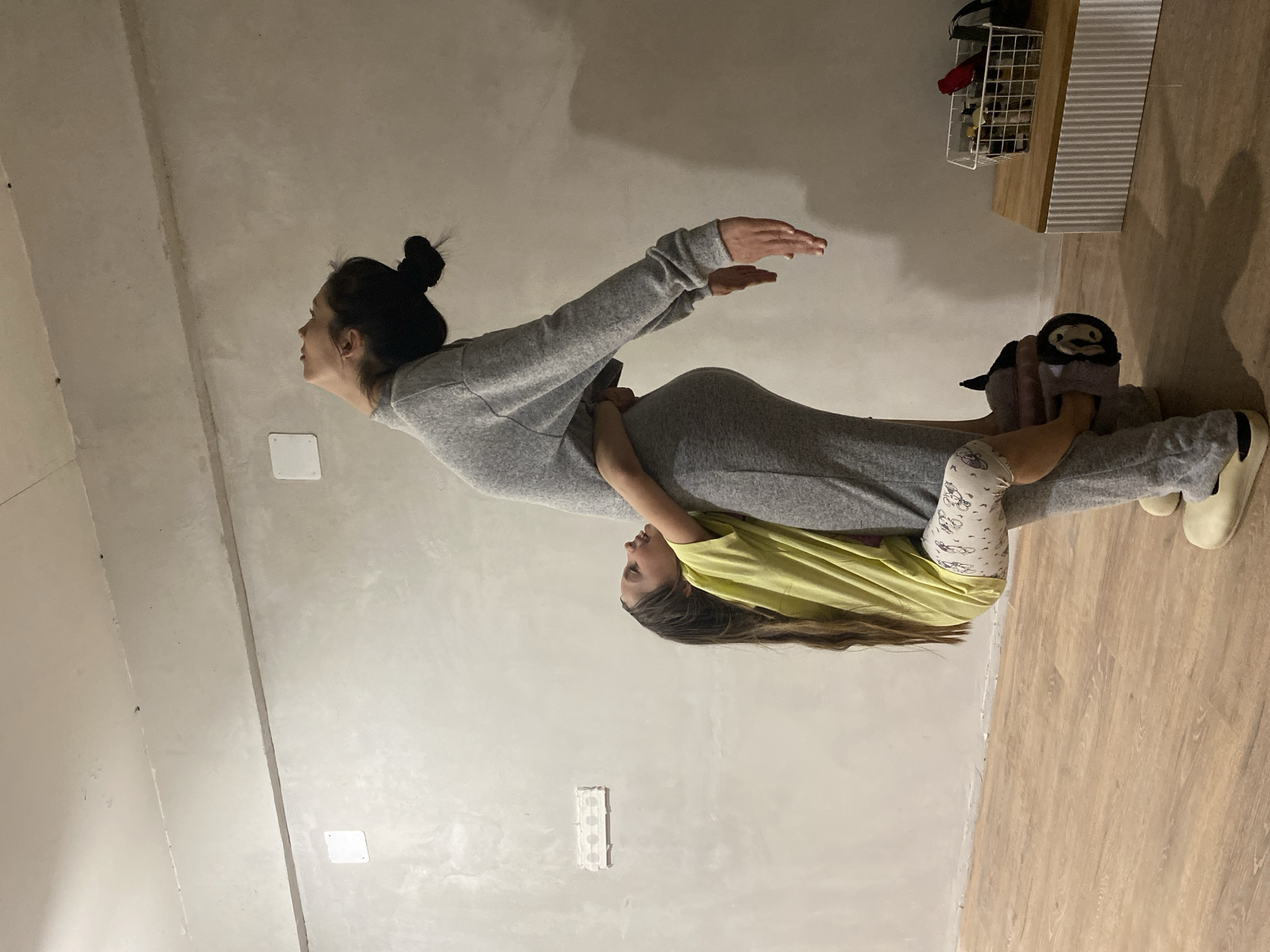 A photo taken in a room in what appears to be a home features a woman with dark hair in a high bun wearing a long grey sweatshirt and grey pants, who appears to be pregnant. She is lying on her back on the floor. A young girl with brown hair wearing a yellow t-shirt is lying across the woman's torso. A plush toy is positioned near the woman's legs. The woman's arms are extended outward. The room has light-colored walls and a wood floor. A wire basket and other objects are visible on a shelf in the upper right corner of the image. There are electrical outlets and other wall fixtures on the wall. This image was not taken in Votkinsk, Russia. The location is undetermined.