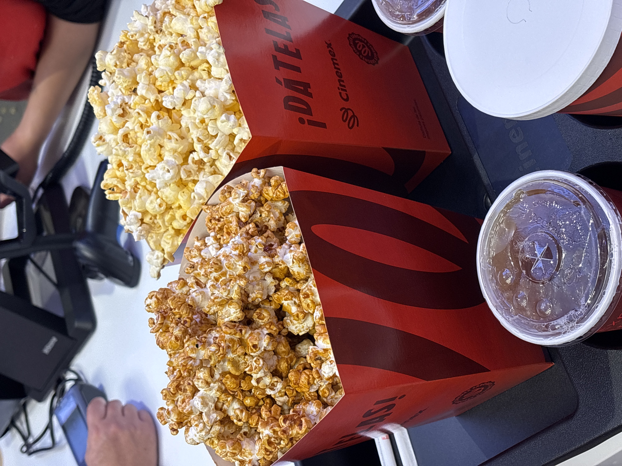 Two large red cartons of popcorn, one caramel-covered and one buttered, along with three covered drink cups are displayed on a black tray. The popcorn cartons have the logo of the Cinepolis movie theatre chain and the phrase "¡Dátelas!" in Spanish. The tray is on a white surface, possibly a table. Part of a person's arm and hand is visible near a black object, possibly a payment terminal. The location is likely inside a Cinepolis movie theater in San Bernardino Tlaxcalancingo, Mexico, based on the theatre chain's logo.