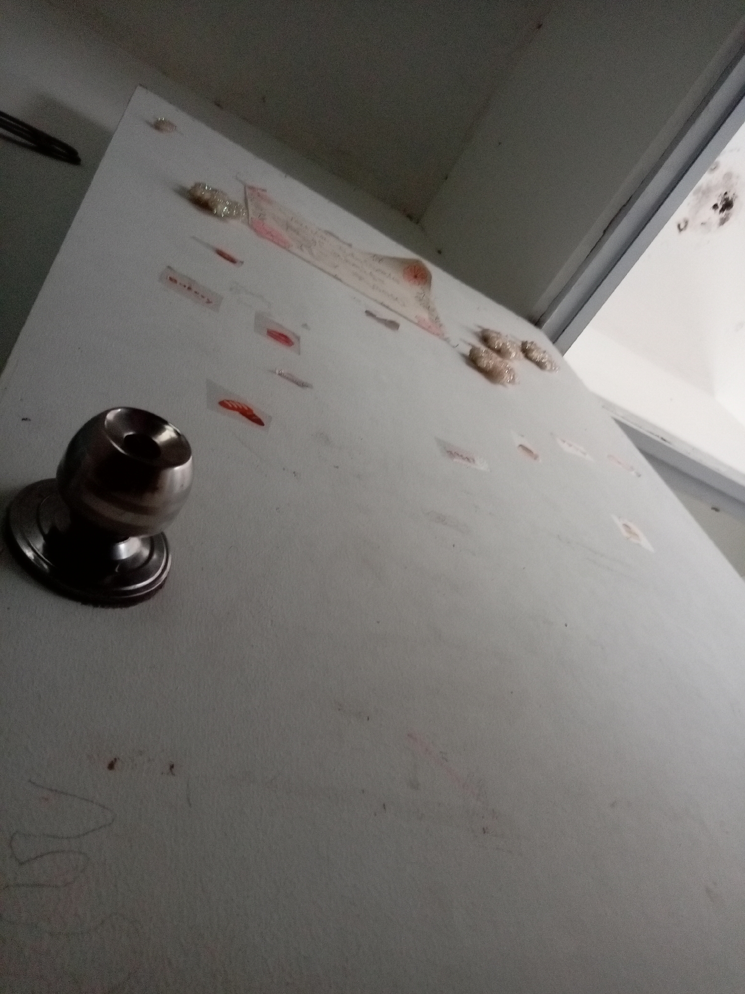 The image shows a close-up, angled view of a white door or cabinet surface. The surface appears to be textured and slightly dirty. A metallic doorknob is prominent in the lower left quadrant. Scattered across the surface are several small, indistinct items, some appearing as smudges or stains, and others as small pieces of paper or tape with writing or images on them. Some of these appear to be red, possibly bloodstains or red ink. There are also some raised, lumpy, light-colored objects adhered to the surface, particularly in the upper right and mid-right areas. In the background, the ceiling or top of the cabinet is visible, with some dark marks or stains on it. The overall impression is that of a used or neglected surface, possibly in an interior space. The image does not provide any specific indicators of its location in Batam, Indonesia.