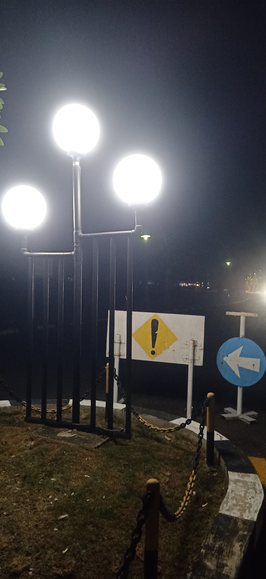 This media file captures a nighttime scene in Tambontunong, Indonesia, featuring a triple-headed lamppost illuminating a dark, outdoor area. The lamppost has three large, spherical, white light fixtures mounted on a dark metal pole, casting a bright glow. In front of the lamppost is a black metal fence or barrier, behind which are two signs. The sign on the left is a white rectangle with a yellow diamond shape in the center, containing a black exclamation mark. The sign on the right is a circular blue sign with a white arrow pointing to the left. The ground is covered in grass and scattered leaves, with a yellow and black chain lying on the ground, appearing to delineate an area or path. The scene suggests a public outdoor space, possibly a park, walkway, or entrance, that is lit for nighttime use. The time of day is clearly night, given the darkness and the artificial lighting. The weather appears to be clear, as there are no visible signs of rain or fog. The overall mood is functional and perhaps a little stark due to the bright lights in the darkness. No people are visible in the image.