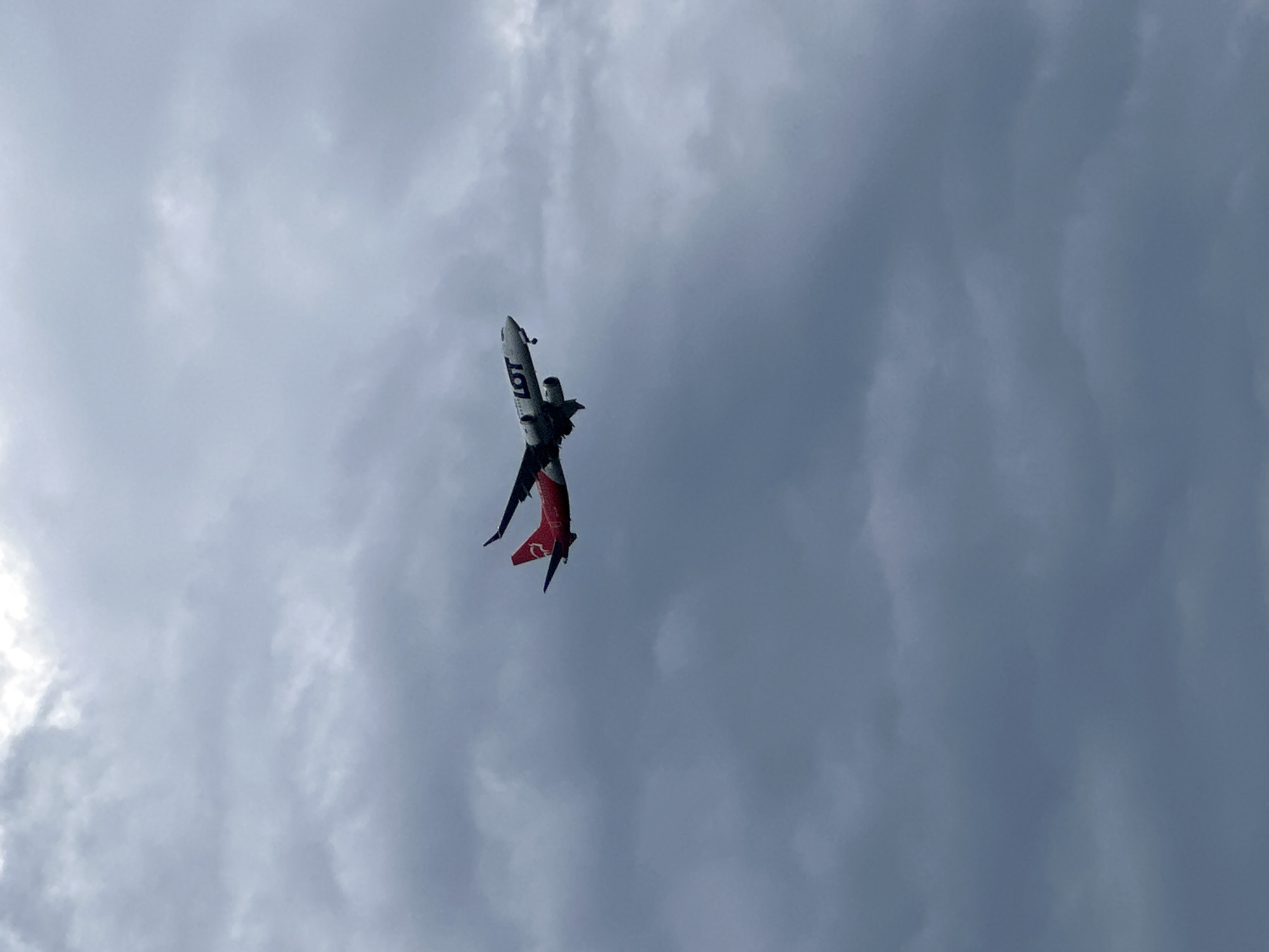 An airplane is captured mid-flight against a heavily overcast and cloudy sky, suggesting a daytime scene with muted light. The aircraft, possibly performing an aerial maneuver, is seen in an inverted or steeply angled position, with its fuselage pointing downwards and slightly to the left from the viewer's perspective. The main body of the airplane is white, and the word "LOT" is clearly visible in dark blue or black letters on its side. The tail section of the aircraft is predominantly red, featuring a white logo or design that resembles the letter "Q". The wings are partially visible, extending from the fuselage. The background is entirely dominated by a dense, grey cloud cover, ranging from lighter shades where light penetrates to darker, more ominous tones. No other objects or ground features are visible, emphasizing the aircraft's isolation in the sky over Burgas, Bulgaria.