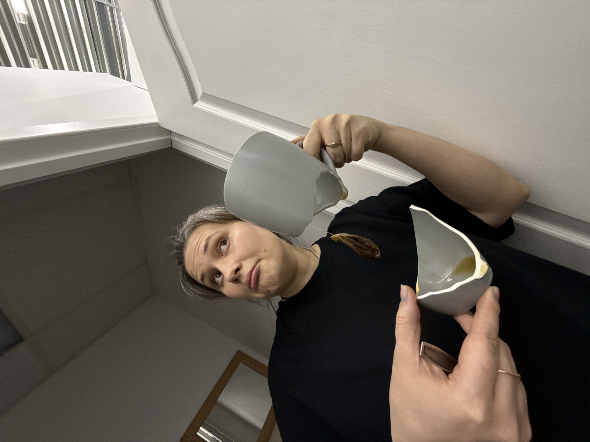An adult female is depicted indoors in Chaikovsky, Russia, holding two broken fragments of a light grey ceramic mug or bowl. She is positioned against a light-colored wall with a wooden-framed mirror visible to her upper left, and a white, paneled door to her right. The woman, exhibiting a surprised or slightly exasperated expression, looks upwards and to her right. Her hair is light grey and appears pulled back. She wears a black short-sleeved top and a necklace with a dark, oval-shaped pendant. Gold-colored rings are present on both her left and right ring fingers, and some fingernails show traces of light-colored polish. She holds the larger, upper portion of the broken vessel in her right hand, near the handle, with an amber-colored liquid (likely a beverage) spilling from the fractured edge. Her left hand cradles the smaller, lower fragment, which also contains a small quantity of the same amber liquid. The ceiling above her is light-colored and textured, with a separate section showing horizontal slats visible through an opening or above the door frame.