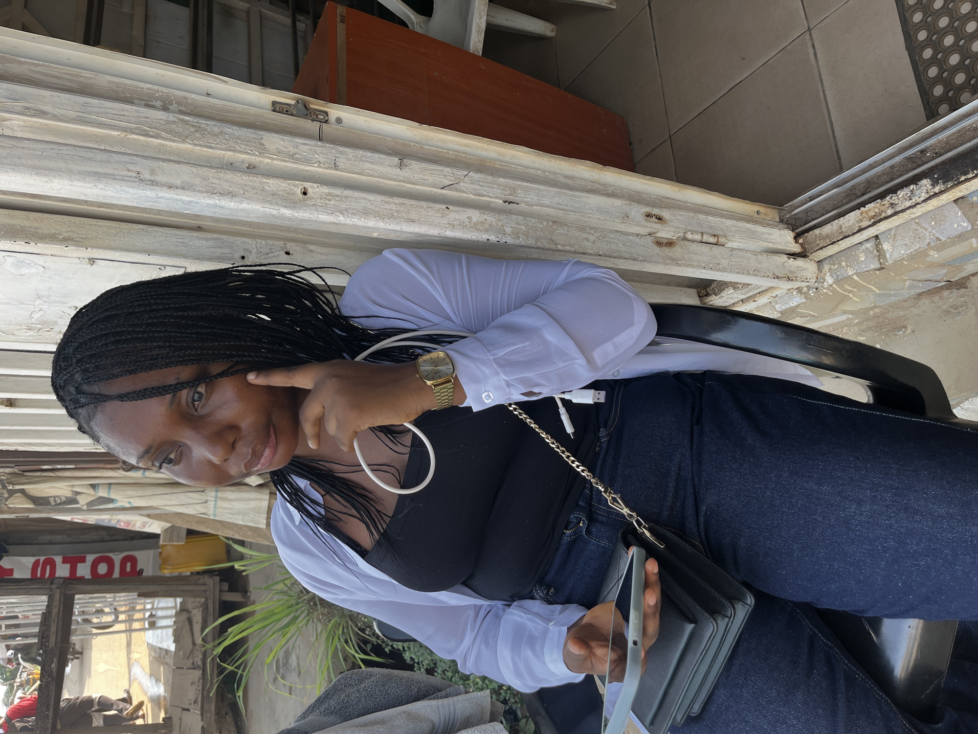 A young Black woman with braided hair is sitting outdoors, holding a tablet in her left hand and resting her right index finger on her cheek. She is wearing a black tank top, a white unbuttoned shirt, and dark blue jeans. A gold wristwatch is visible on her right wrist. She is seated in a black chair, and a black purse with a gold chain strap is slung across her body. The background shows a weathered wooden door or frame, some potted plants, and glimpses of a street scene with people and vehicles, suggesting an urban environment, possibly in Abuja, Nigeria. The overall lighting indicates it is daytime.
