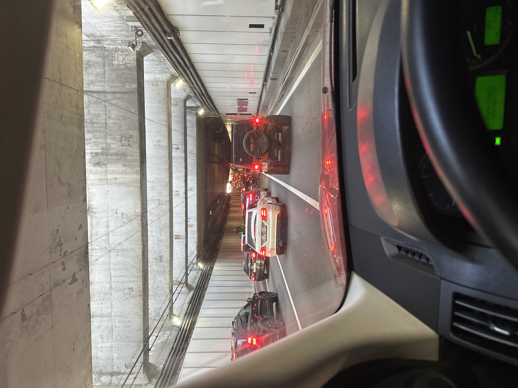 The image depicts a traffic jam inside a tunnel, likely on the M11 Neva Highway near Khimki, Russia, as indicated by the license plates. The perspective is from the driver's seat of a car, showing the dashboard and windshield. Numerous vehicles are visible, including sedans, SUVs, and a Jeep, all stationary or moving slowly. The tunnel structure, with its concrete ceiling and walls, is illuminated by overhead lights, and the traffic lights are red, suggesting a complete stop. The vehicles have their brake lights illuminated, further emphasizing the traffic congestion. The weather appears to be overcast or evening, as the interior lighting of the tunnel is prominent.