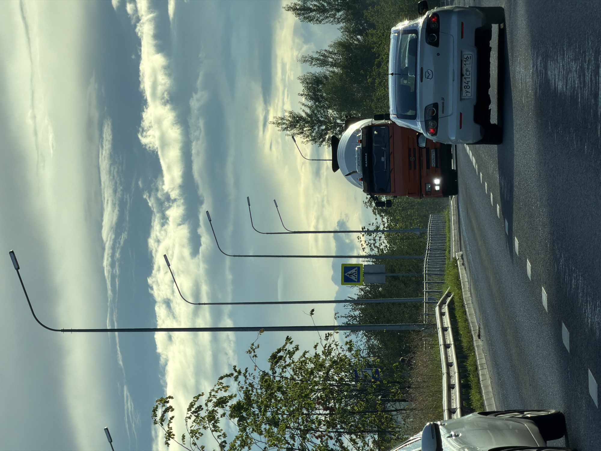 The image captures a view from a moving vehicle on a wet road, likely in Kazan, Russia, given the license plates visible. The sky is overcast with dramatic clouds, casting a diffused light. Several streetlights line the road, some partially obscured by trees. In the foreground, the rear of a white Mazda sedan is visible, its license plate reading "Y841CP 116". Ahead of the Mazda, an orange Kamaz cement truck is in motion, with its headlights illuminated. To the left, part of another vehicle, possibly a silver sedan, is in frame. A pedestrian crossing sign is visible on the right side of the road. Trees and foliage line the sides of the road, with a metal guardrail also present. The road surface appears wet, with reflections visible.