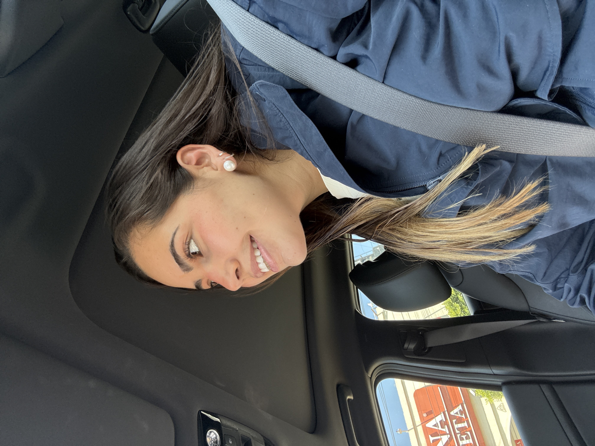 A woman with brown hair and a pearl earring is visible in the front seat of a car. She is wearing a blue jacket and a seatbelt. The interior of the car is black. Through the window, a sign reads "MARIANA ETA" indicating the location is in Puebla, Mexico.