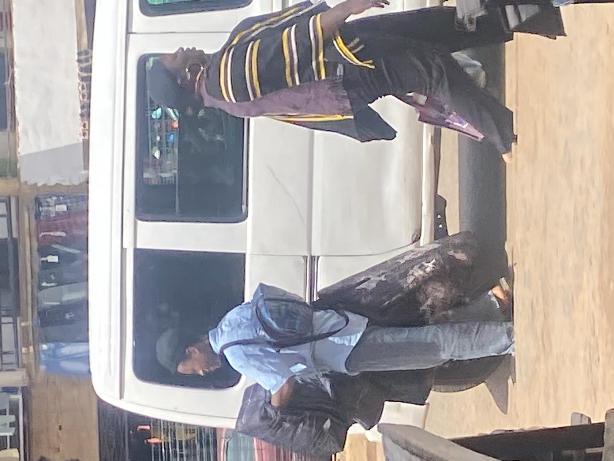 The image shows two people near a white van. The person in the upper portion of the image is leaning out of the van window, wearing a dark top with yellow and white stripes, and purple fabric. Their head is tilted back, and they appear to be wearing earrings. The second person is in the lower portion of the image, also leaning into the van, wearing a light blue collared shirt and dark pants. They have a dark bag slung across their chest and are carrying a large, dark bundle. The scene appears to be outdoors, with a light-colored wall or structure visible to the right of the van. There are reflections in the van windows, suggesting a bright environment. The overall impression is that of a busy, everyday scene, possibly related to transportation or commerce, given the van and the bundles being carried. The orientation of the image is rotated, with the subjects appearing sideways. No visible text is present in the image.