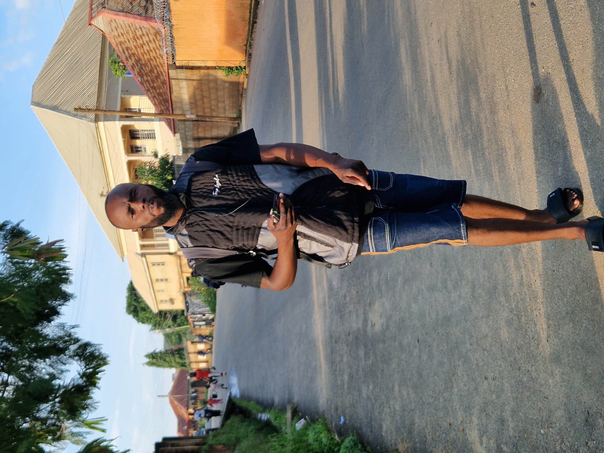 A bald Black man with a beard walks toward the camera on a paved road in an urban area. He wears a black t-shirt with a black and gray vest over it, blue denim shorts with yellow and white stripes on the sides, and black flip-flops. He carries a gray and black backpack and holds a black smartphone in his right hand. In the background, there are several buildings, including a large, light-colored building with a pitched roof and a smaller orange building with a corrugated roof. Other people, including children, are visible walking on the side of the road. Trees and greenery are present on the left side of the image, and the sky is clear and blue. Shadows from the trees are cast across the road. The scene appears to be set in the late afternoon or early evening, given the lighting.
