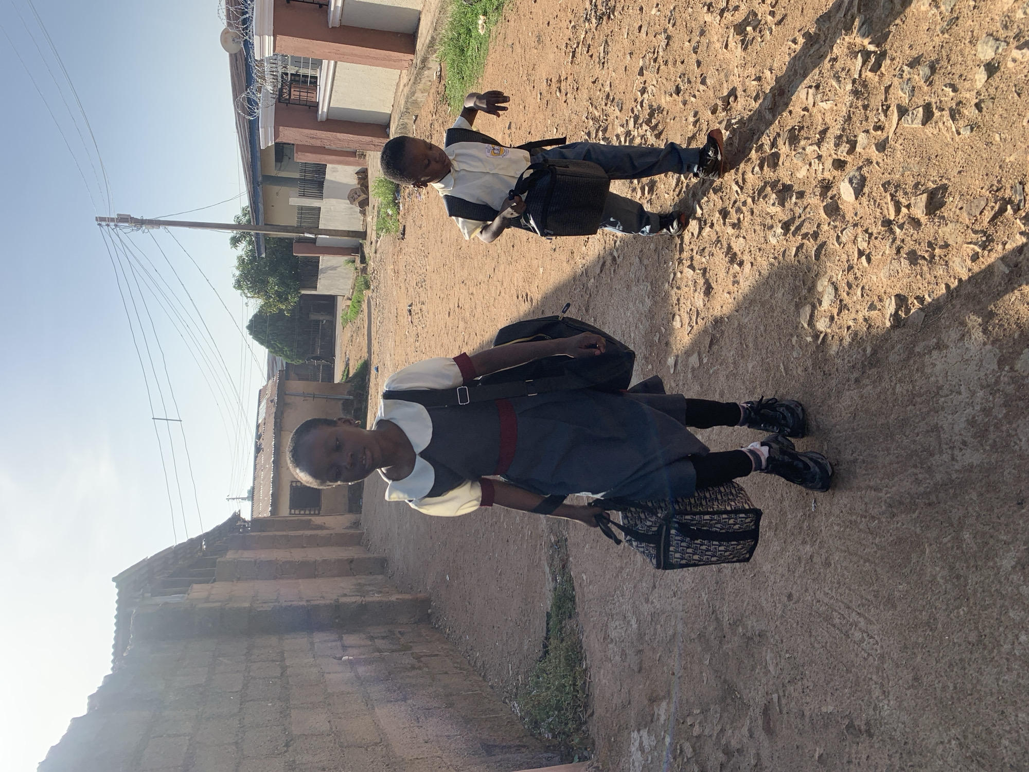 Two school children in uniforms are walking on a dirt road in Gura Top, Nigeria. The first child is a girl wearing a grey dress with a white collar and red trim, black tights, and black shoes. She carries a black backpack and a black and white patterned handbag. The second child is a boy wearing a white shirt with a logo, a grey vest, blue pants, and brown shoes. He also carries a black backpack and a black lunch bag. The road is uneven and strewn with small stones. Buildings of varied construction are visible in the background. An electric pole and wires are present on the left side. The lighting indicates it is daytime.