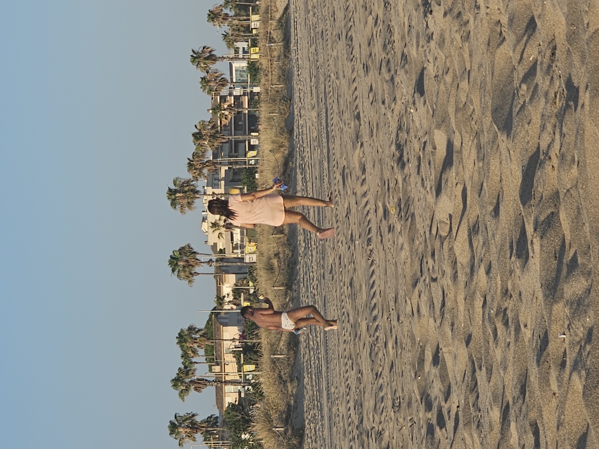 In Castelldefels, Spain, two individuals are walking on a sandy beach. In the foreground, the sand is textured with tire tracks and natural undulations. In the midground, a young person wearing white patterned swim trunks walks towards the left, holding a blue object in their right hand and a light-colored item in their left. To their right and slightly further back, a person wearing a short, pale pink dress walks towards the right, carrying a blue object in their left hand. In the background, a line of palm trees is visible, with buildings and some vegetation behind them. The sky above is clear and blue.