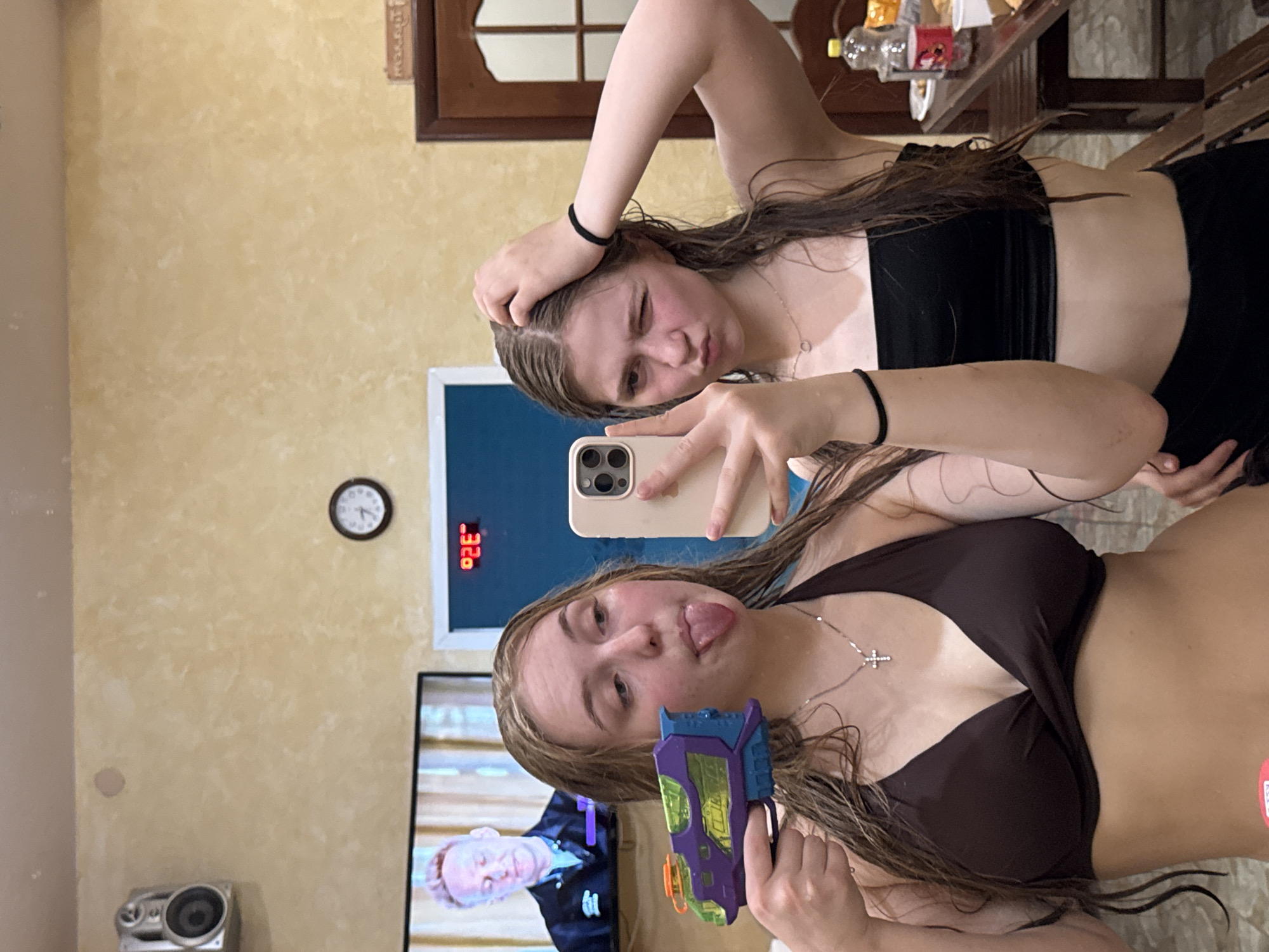 Two young women are taking a selfie. The woman in the foreground is holding a purple and green toy water gun and sticking out her tongue. She is wearing a brown bikini top and a silver cross necklace. The woman in the background is winking and holding her head. She is wearing a black bandeau top. The selfie is being taken indoors, and the background includes a TV screen, a clock, a digital temperature display, a speaker, and a table with various items on it. No specific location within the city of Chaikovsky, Russia can be determined from the image.