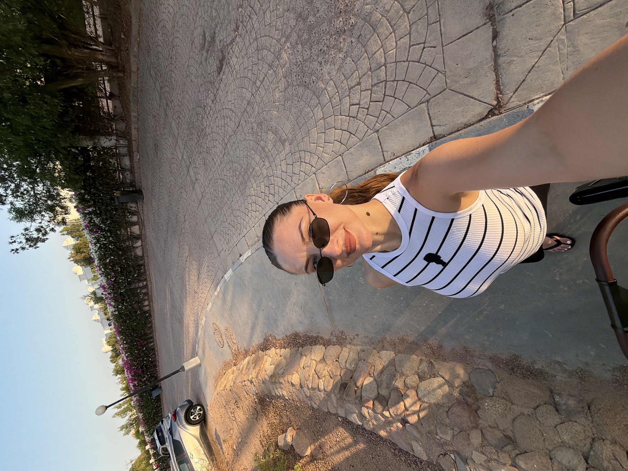 A woman is taking a selfie outdoors in Sharm El Sheikh, Egypt. She is standing on a paved area next to a stone retaining wall. The wall is covered in small, rough stones and separates the paved area from a sandy, dry, arid landscape with some sparse vegetation. Behind her, a line of flowering bushes with pink and white flowers runs along a white wall, with trees and modern white buildings visible in the background. The sun is low in the sky, casting long shadows and highlighting the texture of the paving stones. The woman is wearing a white tank top with black stripes, black sunglasses, and black flip-flops. She has a mole on her left cheek. In the foreground, part of a stroller or pram is visible. A silver car is parked on the paved area to the left of the frame.