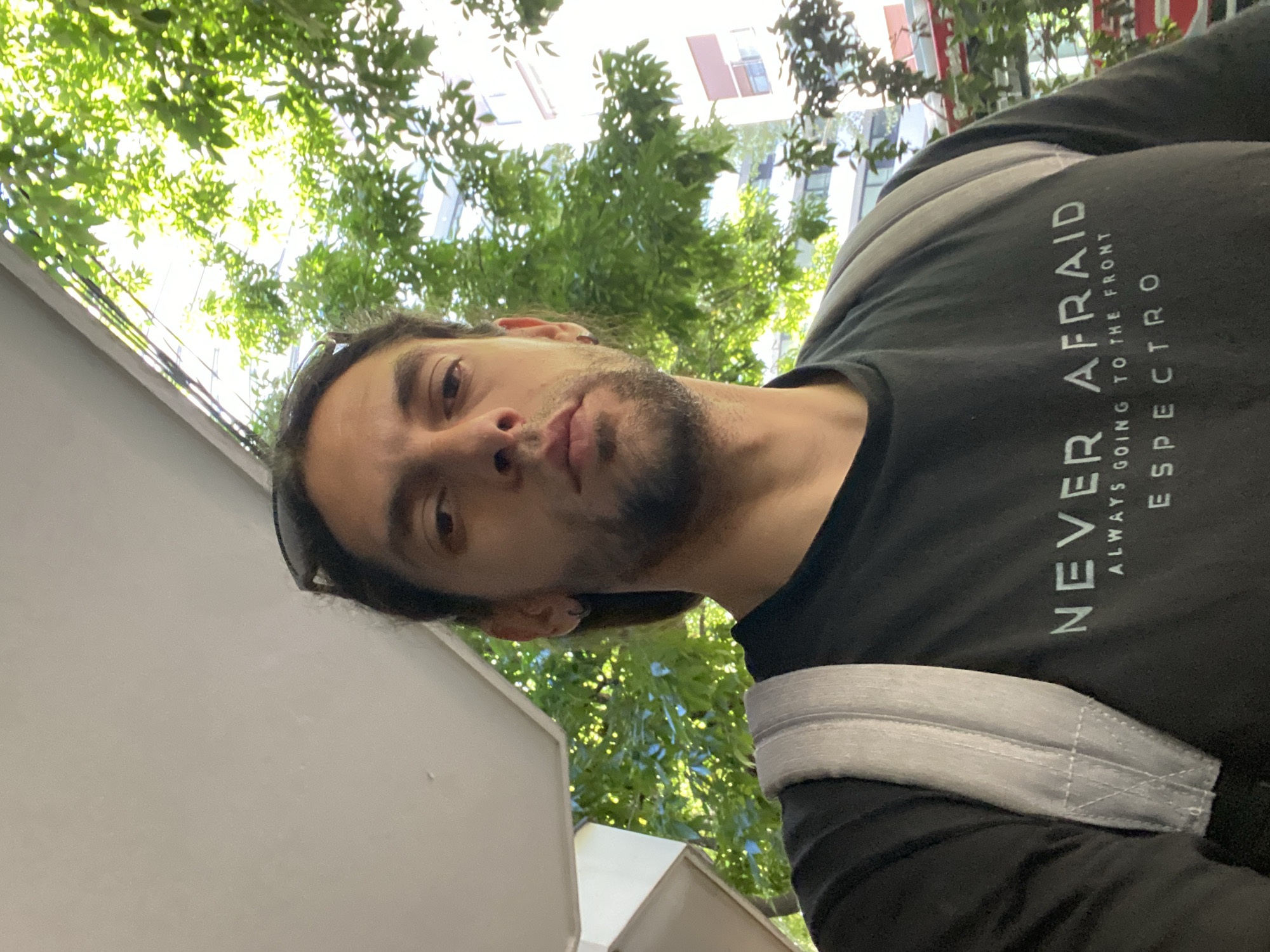 A single adult male is centered in the frame, looking directly forward. He has dark, shoulder-length hair, a light beard, and dark eyes. Sunglasses are perched on top of his head. He wears a small hoop earring in his left ear. He is dressed in a black long-sleeved shirt with white text: "NEVER AFRAID" (large font), "ALWAYS GOING TO THE FRONT" (smaller font), and "ESPECTRO" (medium font). A light gray backpack strap is visible over his right shoulder.
The scene is an outdoor urban environment within the city of Buenos Aires, Argentina. The background features lush green foliage from trees, brightly illuminated by sunlight filtering through the leaves. Partially visible behind the trees are sections of multi-story buildings with light-colored facades, including details such as a dark rectangular window frame and a reddish-brown awning or shutter element. A segment of a light-colored architectural overhang or ceiling is visible in the upper left. The setting is consistent with a tree-lined street or sidewalk in a residential or mixed-use neighborhood common in Buenos Aires, such as Palermo, Belgrano, or Recoleta. The individual is stationary, posing for a photograph.