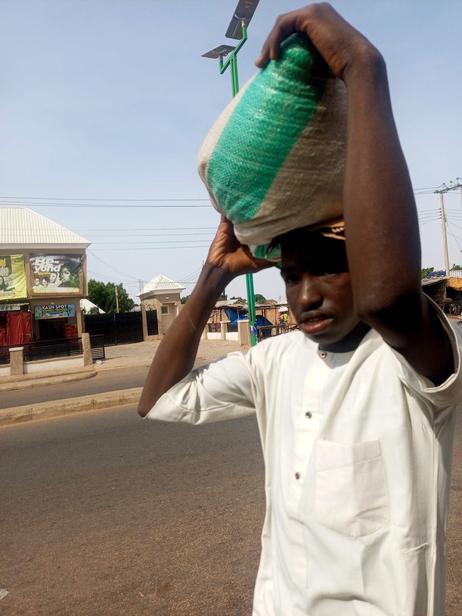 The image depicts a young man in Gombe, Nigeria, carrying a large sack of goods on his head while walking on a paved road. He is wearing a white traditional outfit. In the background, there are buildings, including shops with signage, a street lamp with solar panels, and overhead power lines. The overall setting appears to be an urban environment during the daytime.