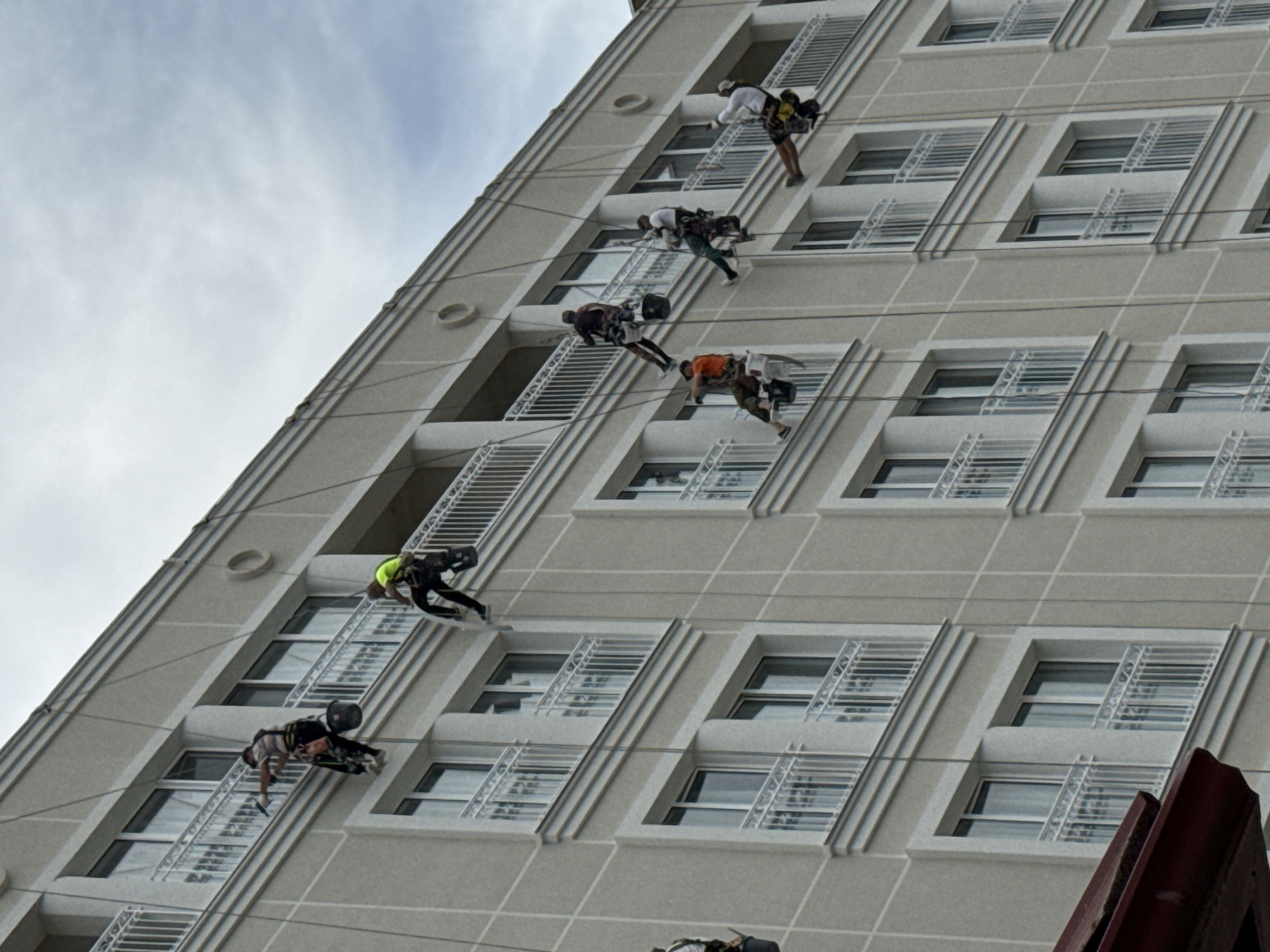 In Kislovodskiy, Russia, a group of individuals are engaged in high-rise window cleaning on the exterior of a multi-story building. The building features a light-colored facade with white balconies and numerous windows. The individuals are suspended by ropes attached to the top of the building, employing specialized equipment for their work. They are equipped with safety harnesses, ropes, and buckets containing cleaning supplies. The workers are positioned at various levels along the building's facade, indicating a systematic approach to cleaning. The sky is overcast, suggesting a potentially cloudy day. The image captures the challenging nature of this profession, highlighting the skill and courage required for such tasks.