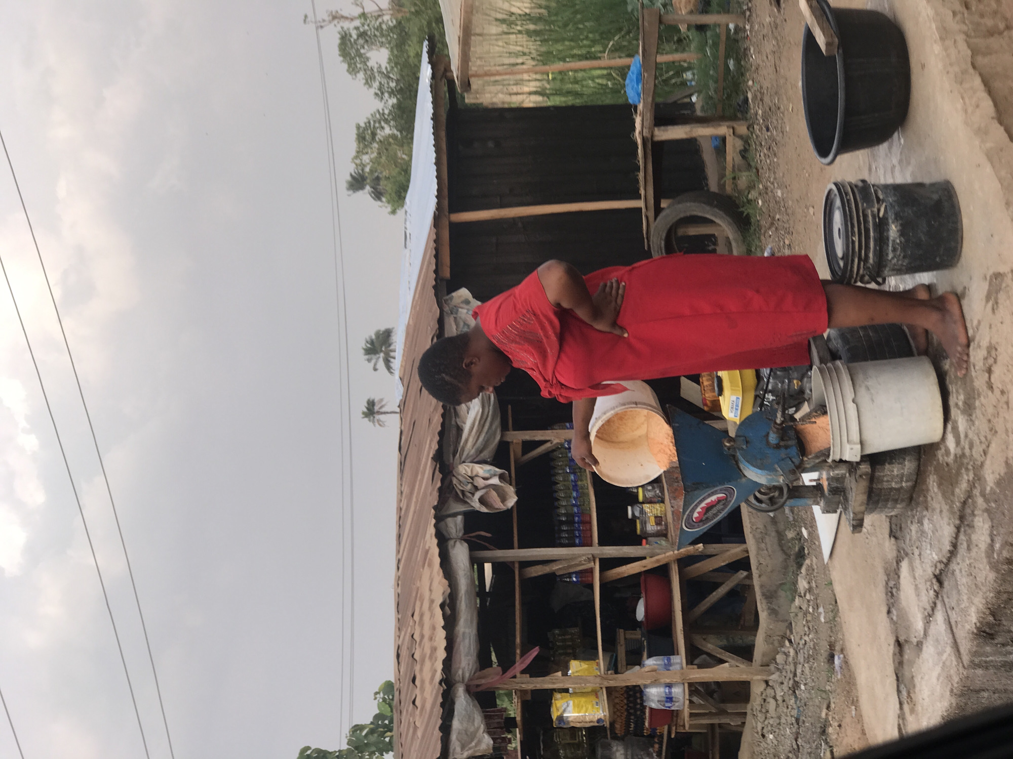 A woman in a red dress is standing next to a grinding machine on a cement surface next to a roadside stall. The stall is constructed of wood and corrugated metal, and contains various products. There are buckets and other containers nearby. The environment appears to be an outdoor, possibly rural, area under an overcast sky. The exact location is Unknown, Unknown.