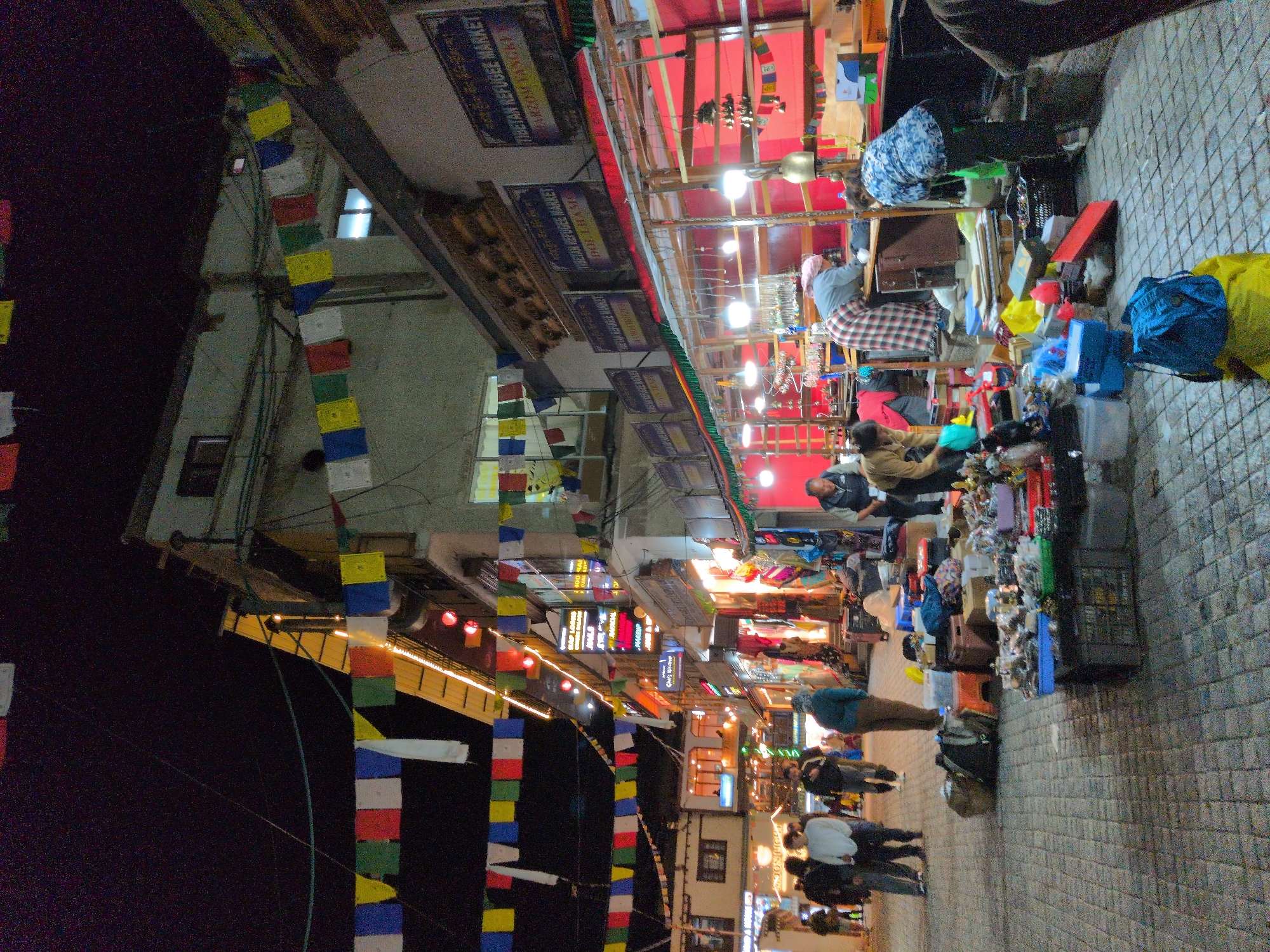 This is a street-level, slightly tilted shot of a bustling market at night in Leh, India. The scene is vibrant with colorful prayer flags strung across the narrow street, fluttering above market stalls laden with goods. Several people are visible, some browsing the merchandise, others walking through the crowded lane, suggesting a lively shopping and social atmosphere.
The market appears to be a Tibetan refugee market, as indicated by signs on the buildings that read "TIBETAN REFUGEE MARKET" and "POLHAM". The stalls are illuminated by bright artificial lights, casting a warm glow on the wares, which include bags, clothing, and various trinkets. The ground is paved with cobblestones.
The time of day is clearly night, evidenced by the darkness in the background and the artificial lighting. The weather appears to be clear. The overall mood is one of energy and commerce, with people engaged in their activities. The presence of prayer flags points to the cultural context of the location. Various objects like bags, shopping baskets, and display racks are prominent.