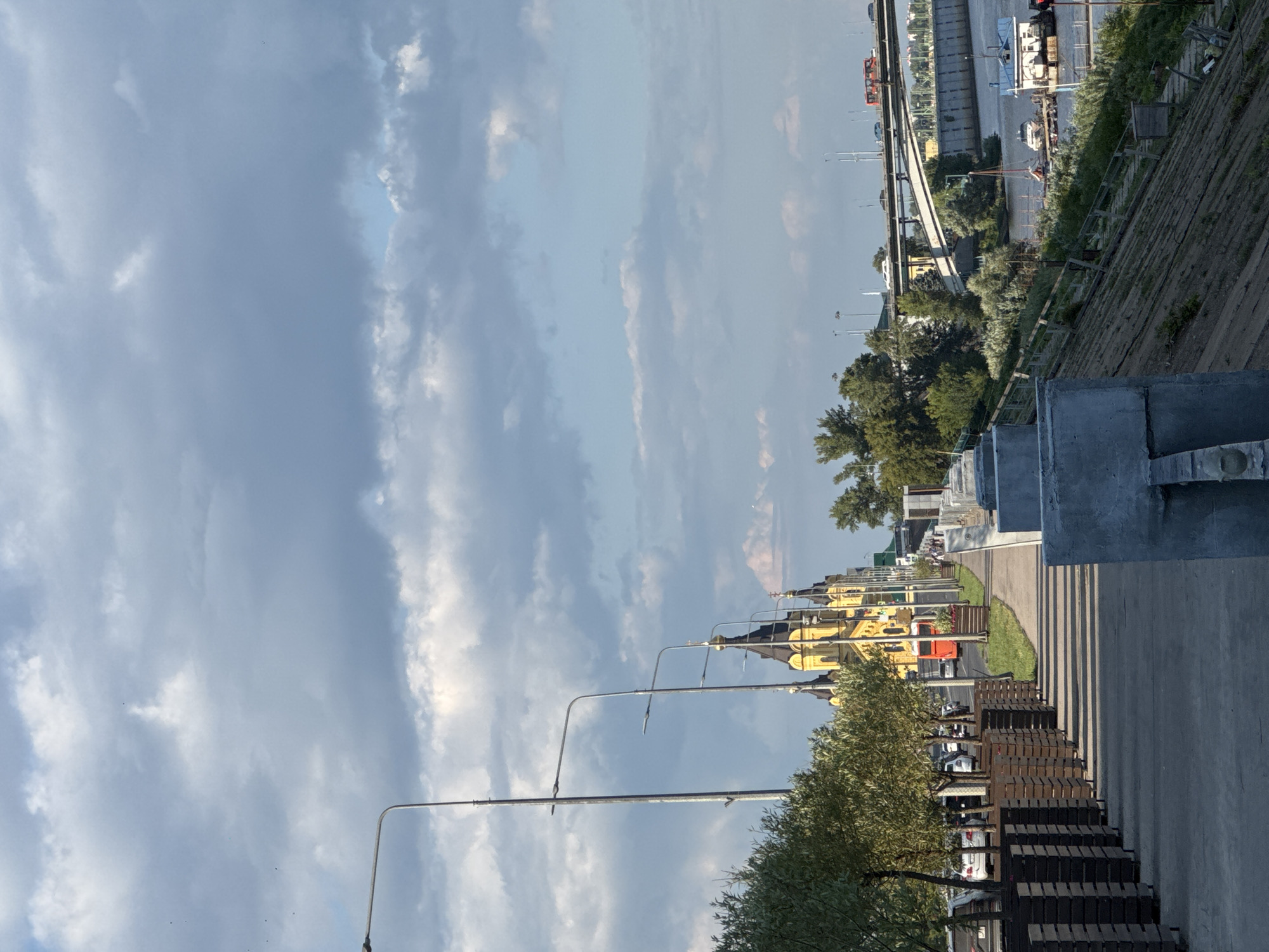 This is a vertical, eye-level, medium shot of a waterfront promenade in Nizhny Novgorod, Russia. The image features a wide, paved walkway with rows of wooden benches and lampposts. Beyond the promenade, a bright yellow building with ornate architectural details, likely a historical landmark or church, is visible. Trees and other greenery line the edge of the walkway, and a street with cars and a red bus runs parallel to it. In the background, a large bridge and a river are visible, with a boat on the water. The sky is partly cloudy with dramatic, wispy clouds, suggesting it could be late afternoon or early evening. The lighting indicates a sunny day with some cloud cover. There is no visible text in the image. The scene appears peaceful and offers a glimpse of urban life by the water.