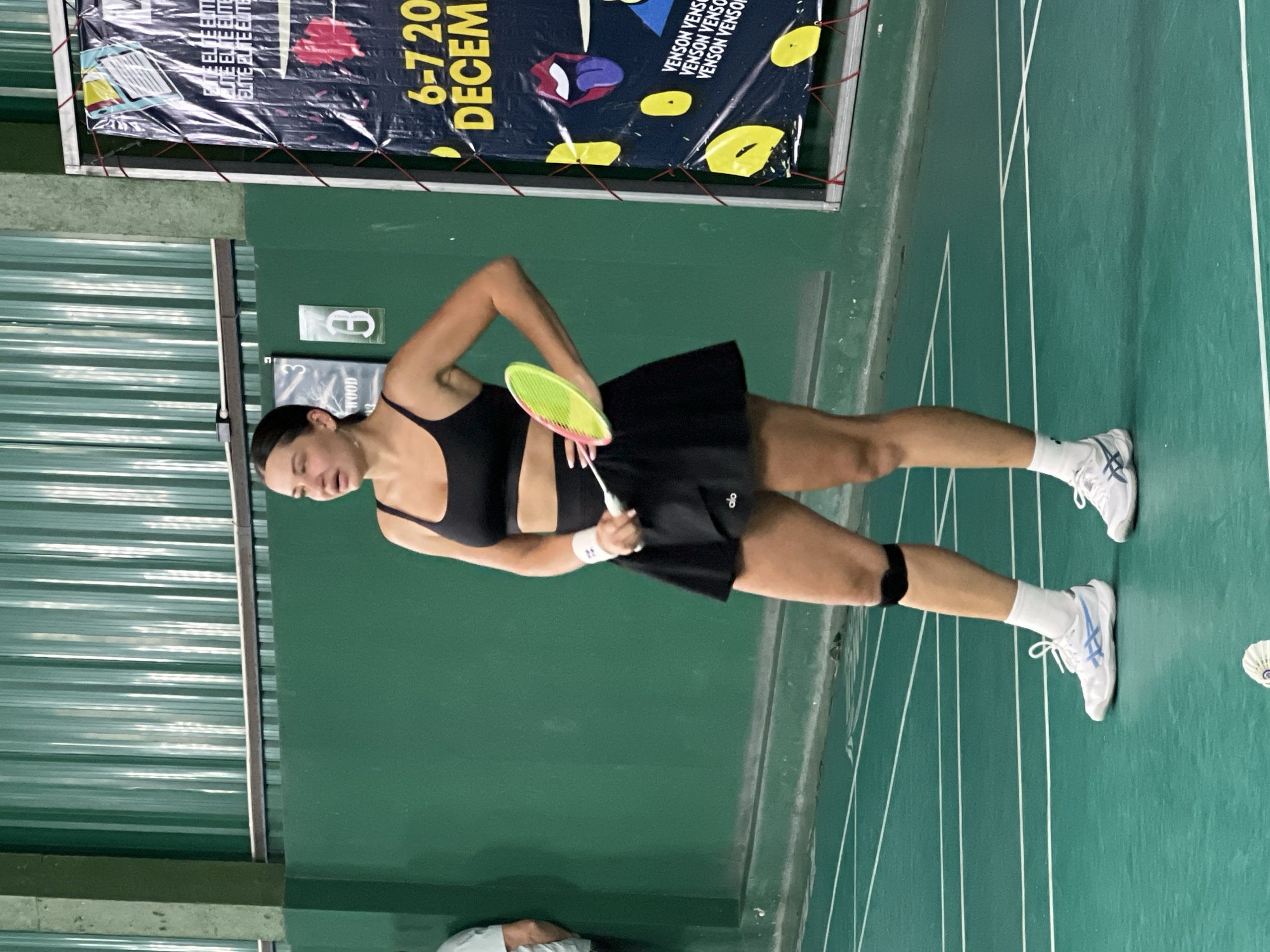 A woman, identified as being in Ban Bang Rak, Thailand, stands on a green badminton court. She is wearing a black sports bra, a black mini-skirt, white athletic socks, and white and blue athletic shoes. A black knee brace is visible on her right knee. She holds a yellow and green badminton racket in her right hand. A white wristband is on her left wrist. In the background, a green wall is visible, with a portion of a banner hanging on the right side. The banner appears to advertise an event with "DECEM" and numbers "6-7 20" visible. The court is marked with white lines.