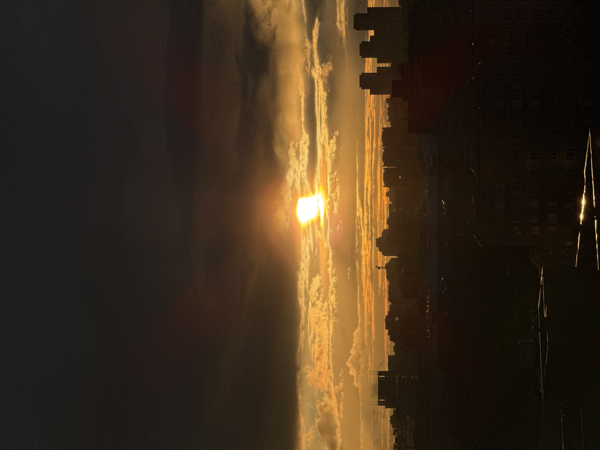The image captures a cityscape at dusk or dawn, likely in Moscow, Russia, given the architectural style of the buildings. The sun is positioned in the center, partially obscured by clouds, creating a dramatic lighting effect with golden hues. Tall buildings form a silhouette against the bright sky. The left side of the image is dominated by dark shadows, contrasting with the illuminated cityscape. Power lines are visible in the foreground on the right. There are no people visible. The overall scene conveys a sense of urban tranquility under a vibrant sky.