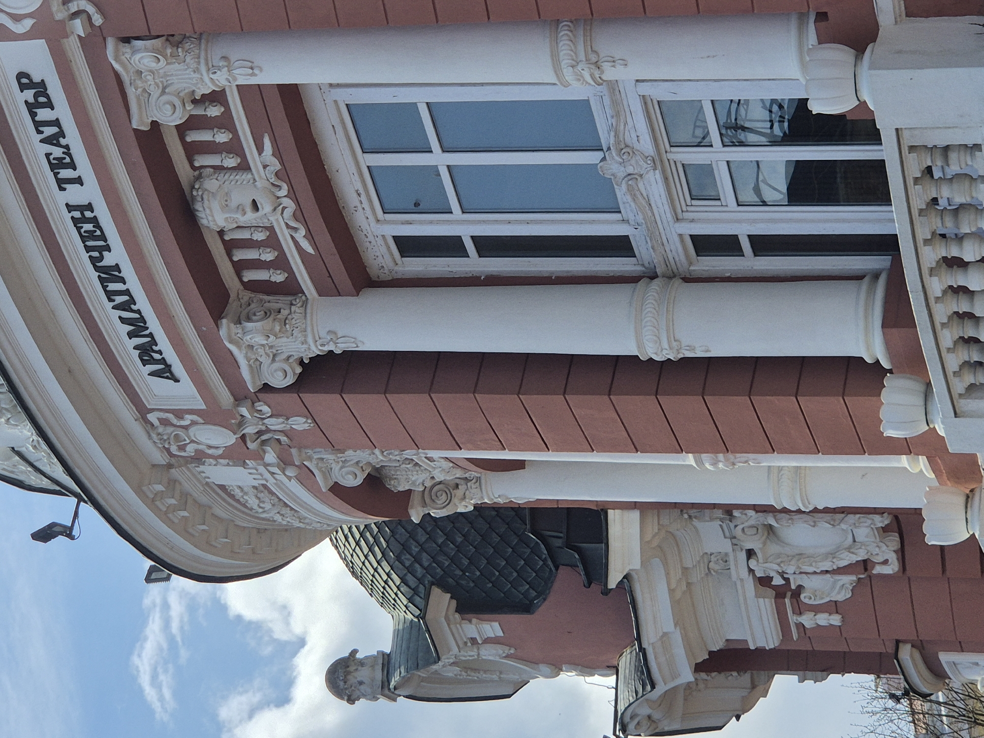 A low-angle shot features a segment of the facade of the "ДРАМАТИЧЕН ТЕАТЪР" (Dramatic Theatre) building in Unknown, Unknown, Bulgaria. Architectural details visible include white columns with decorative capitals, a window with multiple panes, and ornamental moldings. The building’s exterior is painted in shades of white and reddish-brown. Portions of a dome-like structure and statuary are visible at the bottom of the frame. The sky is visible in the lower left corner.