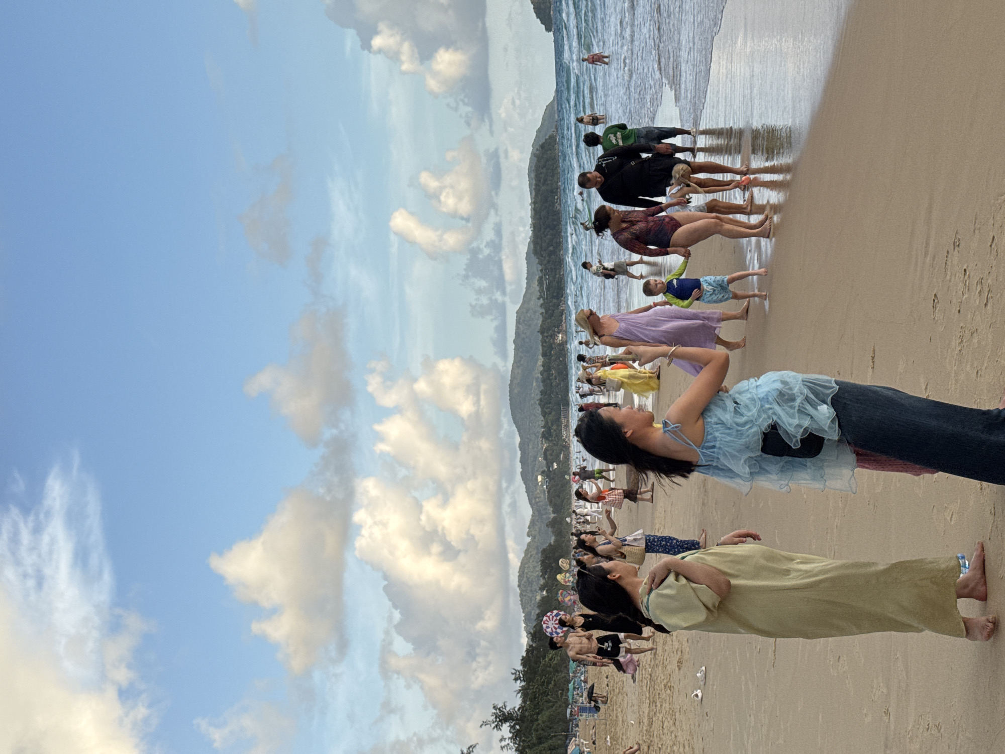 A wide shot captures Karon Beach in Karon, Thailand, under a partly cloudy sky. A diverse group of people strolls along the sandy shore and wades in the ocean. In the foreground, two women stand facing away from the camera, one in a light green, long-sleeved dress and the other in a blue ruffled top and dark pants. To their right, a family walks towards the water; a woman in a patterned swimsuit holds hands with a young boy in a blue and yellow rash guard, followed by a man in a black shirt and a child with blonde hair. Further down the beach, several individuals are in the shallow water. The background reveals lush green hills bordering the coastline, with a few buildings visible in the distance. The sea is a vibrant blue with gentle waves.