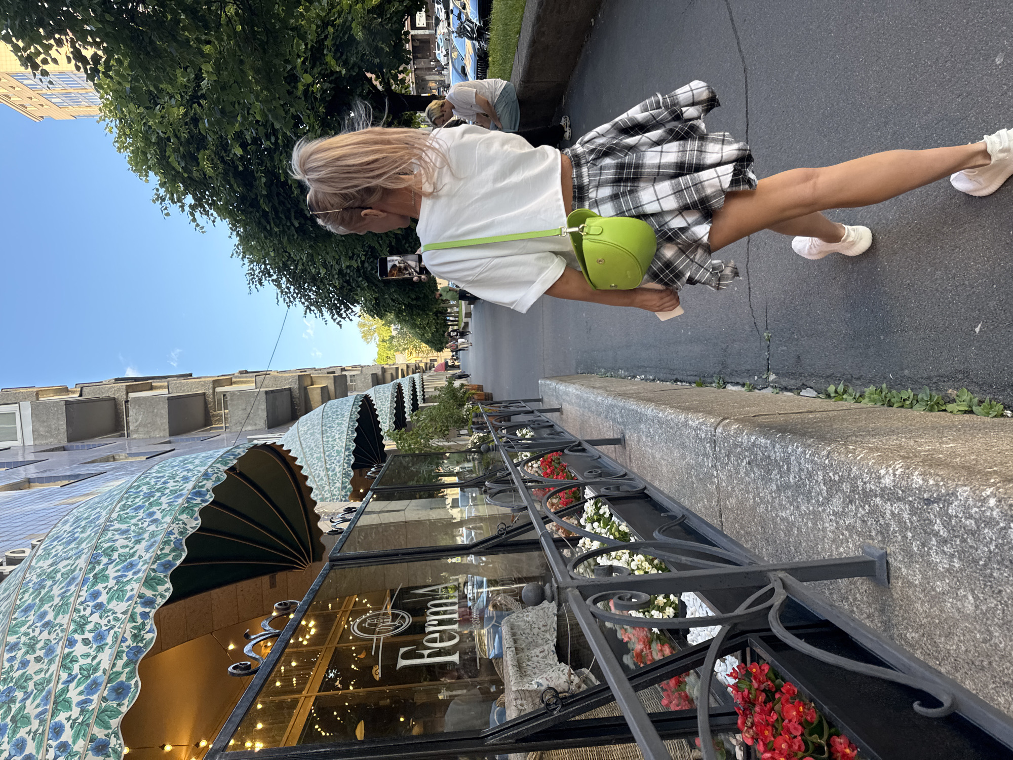 The image depicts a woman walking on a sidewalk in Saint Petersburg, Russia, with a building and trees flanking the street. She is wearing a white t-shirt, a plaid skirt, and light-colored sneakers, and is carrying a bright green purse and a smartphone. The building on the left has awnings with a floral pattern and glass storefronts. A black metal fence with flower boxes is in the foreground. Further down the street, another person is seated on a low wall. Cars are parked along the opposite side of the street, and trees line the sidewalk. The sky is clear and blue, suggesting a daytime scene.