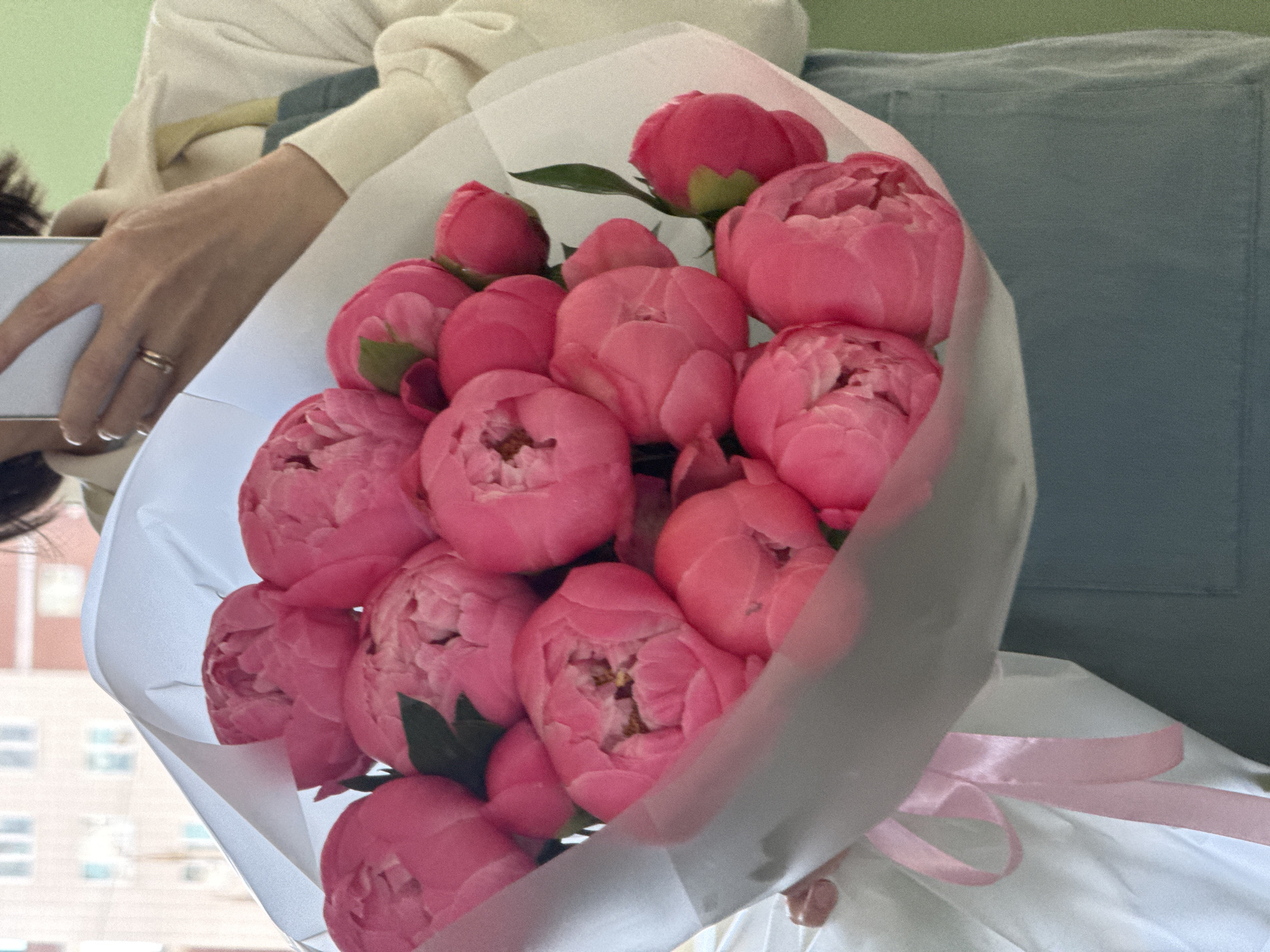 A person holds a bouquet of pink peonies wrapped in white paper with a pink ribbon. A hand with a ring is visible holding a white phone. The background shows an apartment with a blue cushion and a window overlooking a building. It is not possible to determine the location is Izhevsk, Russia based on visual information.
