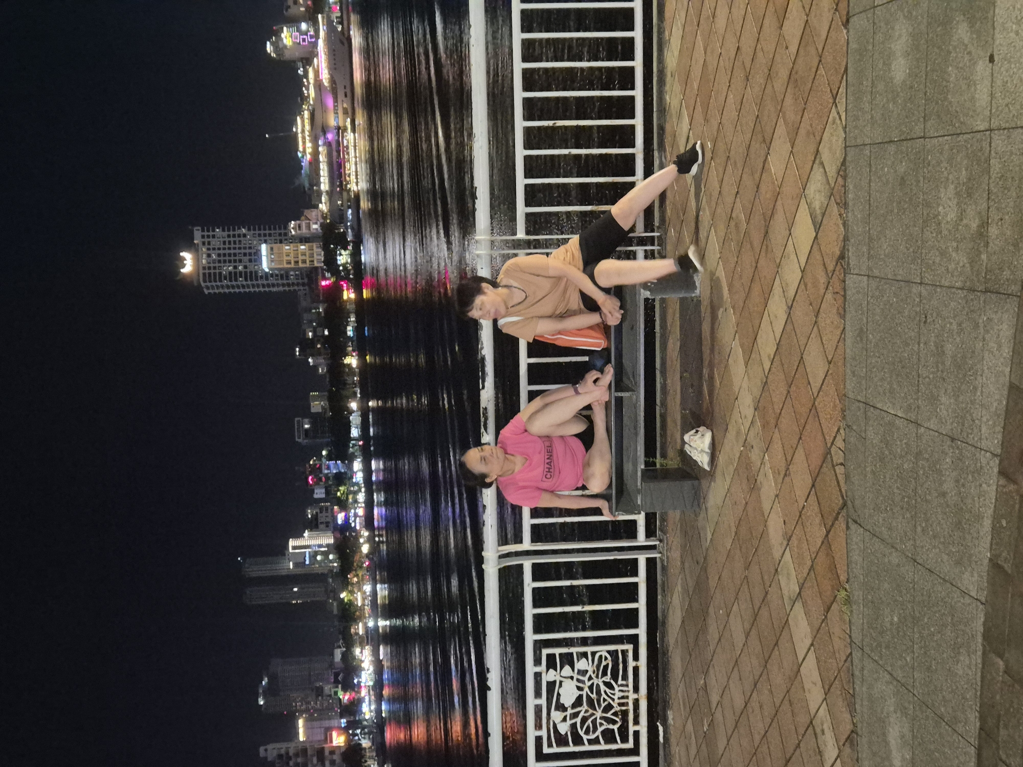 The image captures a nighttime scene along the Han River in Da Nang, Vietnam. Two women are seated on a dark-colored bench facing the water. The woman on the left is wearing a bright pink t-shirt with the word "CHANEL" printed on it and black shorts. She is sitting with her legs crossed. The woman on the right is wearing a tan t-shirt and black shorts. She is leaning back on the bench with her legs extended. A bright orange bag is placed next to her.
In the background, the Han River reflects the lights of the city. Various buildings, including high-rises and a large cruise ship or boat, are illuminated along the waterfront. The sky is dark, indicating it is night. The foreground consists of a tiled walkway, with a white metal railing separating the walkway from the bench area. A white plastic bag is visible on the ground near the bench.