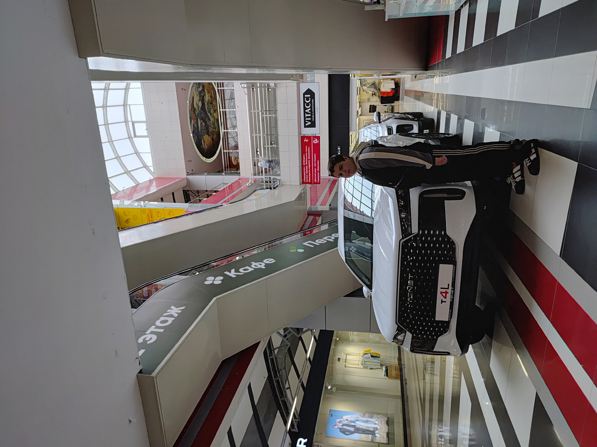 Here is a detailed description of the image:
**Scene Overview:**
The image captures an indoor setting, possibly a shopping mall or commercial center. The perspective is slightly skewed, tilting to the right.
**Environment:**
The setting is modern with elements such as escalators and large windows. The floor is patterned with alternating black and white stripes, with a section featuring a red stripe. Signage in Russian is visible, including a sign indicating "Kafe" (Cafe) and "Etagh" (Floor). There are also various storefronts and advertisements.
**People:**
A male individual stands near the center of the image. He appears to be wearing a jacket and black athletic pants with stripes. His attention seems directed toward the camera or something to his right.
**Objects:**
The dominant object is a white vehicle prominently displayed with the brand "TENET" and model "T4L" indicated on its front. Other notable objects include escalators, storefront signs such as "VITACCI," and advertisements.
**Actions and Interactions:**
The male individual is standing near the vehicle, potentially observing it or posing for the photo. The scene is indoors, likely within a shopping mall or exhibition space.
**Location:**
Based on the OCR text (Optical Character Recognition) and signs present, the location may be in Izhevsk, Russia. The signage includes brand names like "VITACCI", as well as the word "Etagh" (Floor), which is a Russian loanword for the French word "étage". This is indicative of the Russian location.