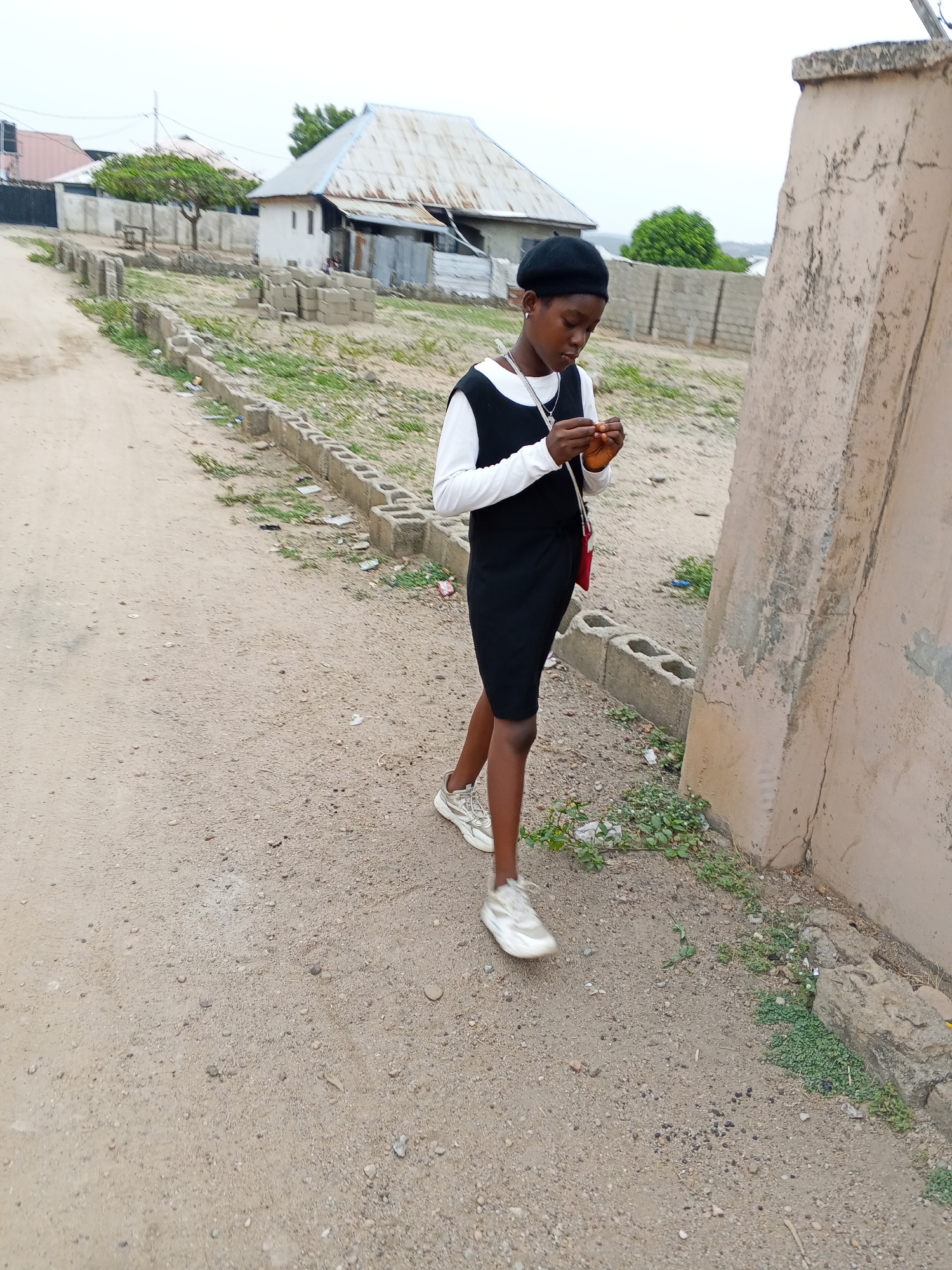 A young woman stands on a dirt road in Adonkolo, Nigeria. She wears a black beret, a long-sleeved white shirt under a black dress, and white sneakers. A small red bag hangs from her shoulder. She is looking down at something she holds in her hands. The road is bordered by concrete blocks and a tan wall. In the background, there are houses with corrugated metal roofs and some trees. The sky is overcast.