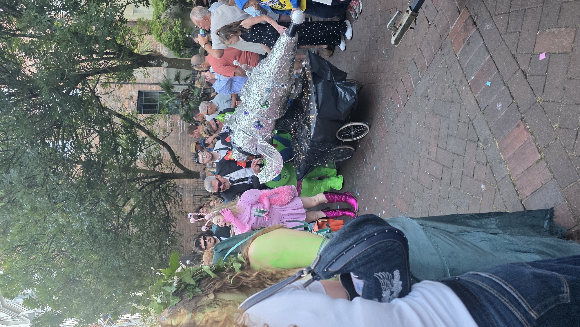A street parade is taking place on a brick-paved street in St Leonards, United Kingdom. A person in a pink polka dot dress and pink boots, with pink feathery accessories, stands near the center of the image. To their right, a large, silver, foil-covered object resembling an alien or fantastical creature is positioned on a black wheeled cart adorned with fairy lights. Another person is dressed in a full green alien costume, partially obscured by the silver creature. Several other people, some in formal attire like suits, are observing the parade. In the foreground, the bottom portion of a person's blue jeans and a dark denim bag are visible. The environment includes trees with green foliage and brick buildings in the background. Confetti is scattered on the ground.