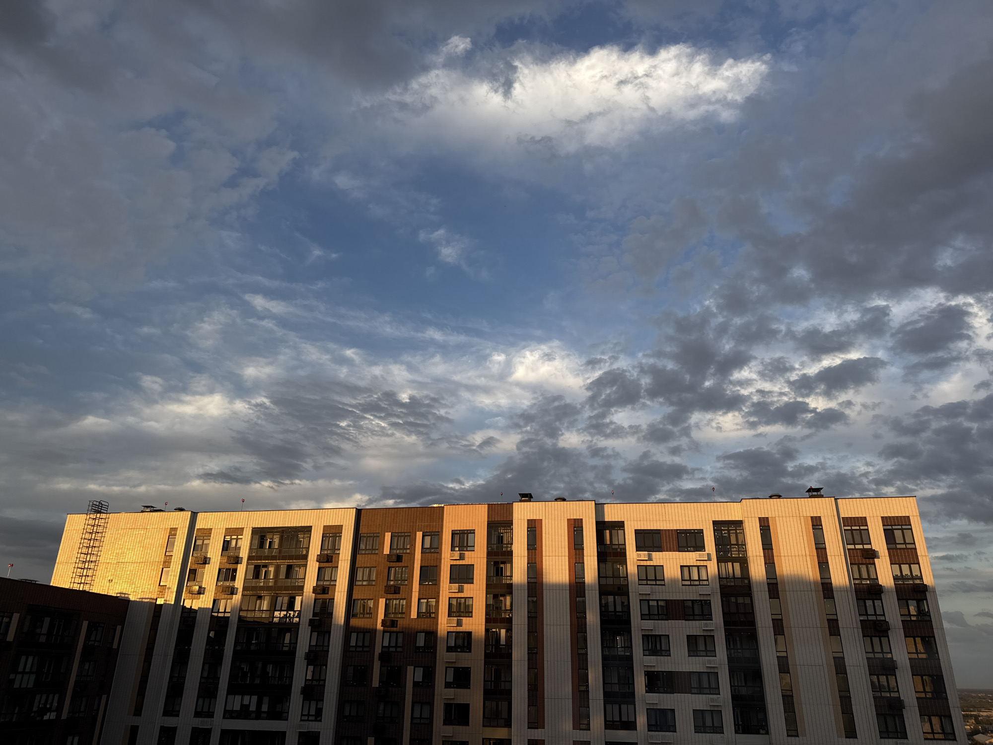 The photograph captures a modern, multi-story apartment building in Lyubertsy, Russia, under a dramatic sky filled with clouds. The building features a varied facade, with sections clad in light-colored tiles and others in darker materials, interspersed with numerous windows and air conditioning units. The setting sun casts long shadows across the building's surface, highlighting the architectural details and creating a striking contrast between light and dark. The sky above is a dynamic mix of blue patches and heavy, gray clouds, some illuminated by the warm glow of the sunset. A fire escape ladder is visible on the left side of the building. The overall scene conveys a sense of urban living during a dynamic weather event.