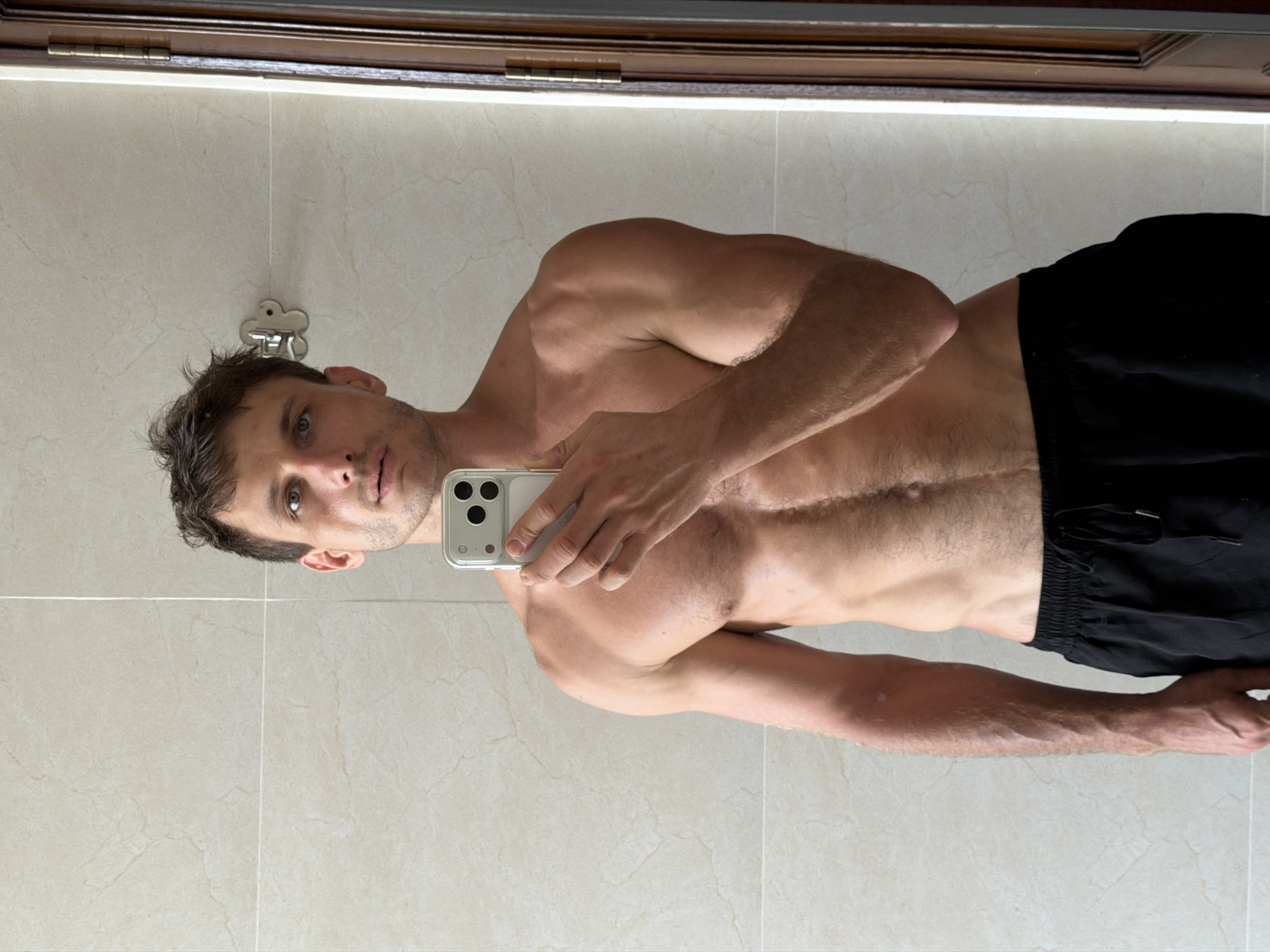 The image is a mirror selfie featuring a shirtless man, captured indoors, likely in a bathroom or changing area within Da Nang, Vietnam. The subject appears to be a man in his 20s or 30s, with short, dark hair, light-colored eyes, and some facial stubble. He maintains a neutral, direct gaze towards the camera.
He possesses a muscular and athletic physique, with well-defined shoulders, arms, and torso. A prominent, linear scar runs vertically down his upper abdomen/lower chest. The man is wearing black shorts and is holding a light-colored smartphone, possibly an iPhone, in his left hand to take the photograph. The phone's distinctive multi-lens camera array is visible.
The background, reflected in the mirror, consists of light-toned, possibly beige or off-white, ceramic or porcelain tiles with visible grout lines, suggesting a clean and modern interior. Above the tiled section, a dark wooden frame or cabinet is reflected. Behind the man's head, a small, metallic, cloud-shaped hook or decorative fixture is visible in the reflection. The lighting suggests an indoor setting, likely with artificial illumination, making the time of day and external weather conditions indiscernible. No legible text is visible within the image.
