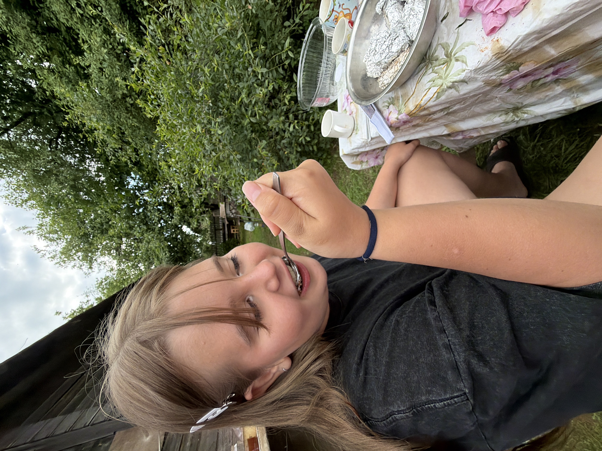 This media file shows a young girl eating from a spoon outdoors in a backyard setting in Бронница, Russia. The girl is lying down, with her head tilted upwards and her eyes closed as she enjoys her food. She is wearing a black t-shirt and dark jeans. Her hair is long and light brown, held back by a white clip. A dark blue bracelet is visible on her wrist.
In the background, a picnic table is set with a floral tablecloth. On the table are various items, including a clear bowl, a metal bowl covered in aluminum foil, a few white cups, and some pink napkins. To the left of the girl, there is lush green foliage and a cloudy sky can be seen. The overall impression is of a casual outdoor meal on a possibly warm day, with a focus on the girl's enjoyment of her food. No visible text is present in the image.