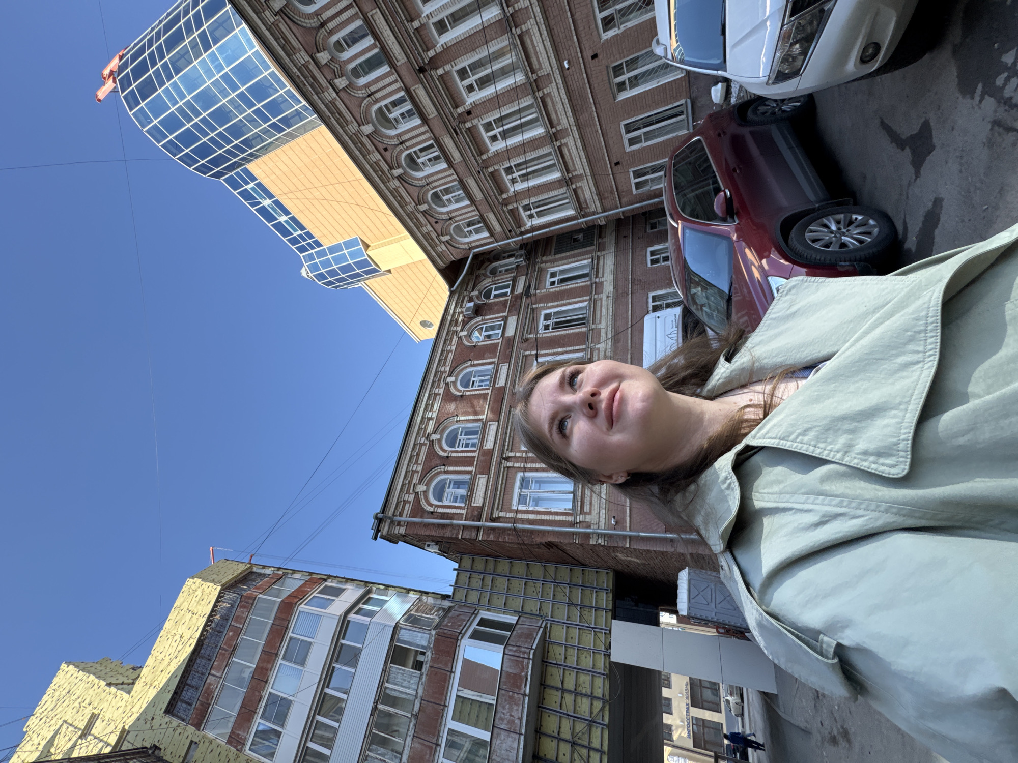 A young woman in a light green jacket stands on a street in Perm, Russia, looking up and slightly to the left. Buildings surround her, including a tall brick building with arched windows, a modern glass and metal structure, and an older building undergoing renovation. Two cars, one white and one dark red, are parked nearby. The sky is clear and blue.