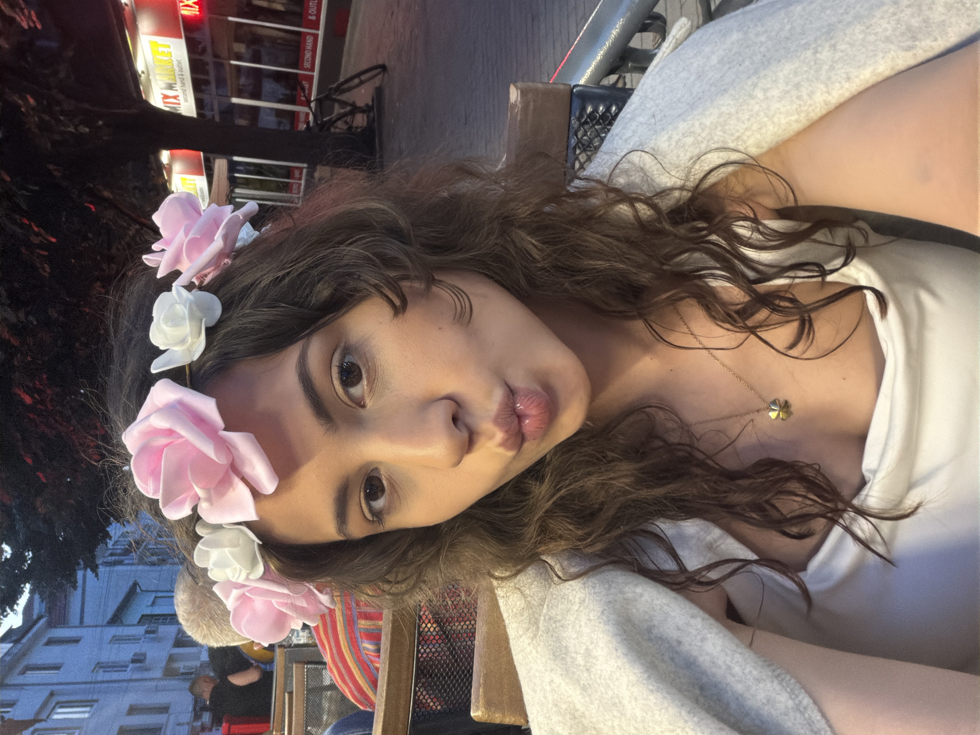 A young adult female is centered in a close-up, self-portrait style image, likely taken outdoors in an urban environment during dusk or early evening. She has medium-dark brown, curly hair and is wearing a floral headband adorned with large pink and white faux roses. Her eyes are brown, and she has a pouting expression with her lips pursed. She is wearing a white top with a draped neckline and has a light grey, textured blanket or cardigan draped over her shoulders. A thin gold chain necklace with a four-leaf clover pendant is visible on her chest.
She is seated at an outdoor cafe or restaurant, with parts of a wooden chair and a black mesh panel visible behind her. To her left, behind her shoulder, the back of a light-skinned person's head with very short or bald hair is visible, along with another person wearing a red and white striped garment. Further back, multi-story buildings with light-colored facades and air conditioning units are visible, indicative of a street scene.
To her right, a dark tree trunk and sparse foliage are present. Behind the tree, a commercial building with illuminated red and yellow signs is visible. The primary sign reads "MIX MARKET" with "second hand & outlet" in English below it. A bicycle is parked near the building. The ground appears to be a paved street or sidewalk. The lighting suggests a transition from daylight to night, with some artificial lights already prominent. The location is Unknown, Unknown.
